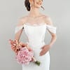 Eine lächelnde Braut im schulterfreien weißen Hochzeitskleid S26-F09 hält einen Strauß rosafarbener Blumen und blickt zur Seite vor einem schlichten hellen Hintergrund.