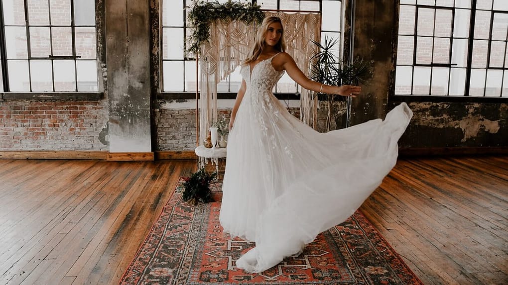 Eine Braut in einem eleganten Hochzeitskleid posiert auf einem Vintage-Teppich. Hinter ihr: rustikale Backsteinwände, große Fenster und Gründekor. Der Raum mit Holzboden verkörpert stilvolle Industrial-Chic Brautmoden.