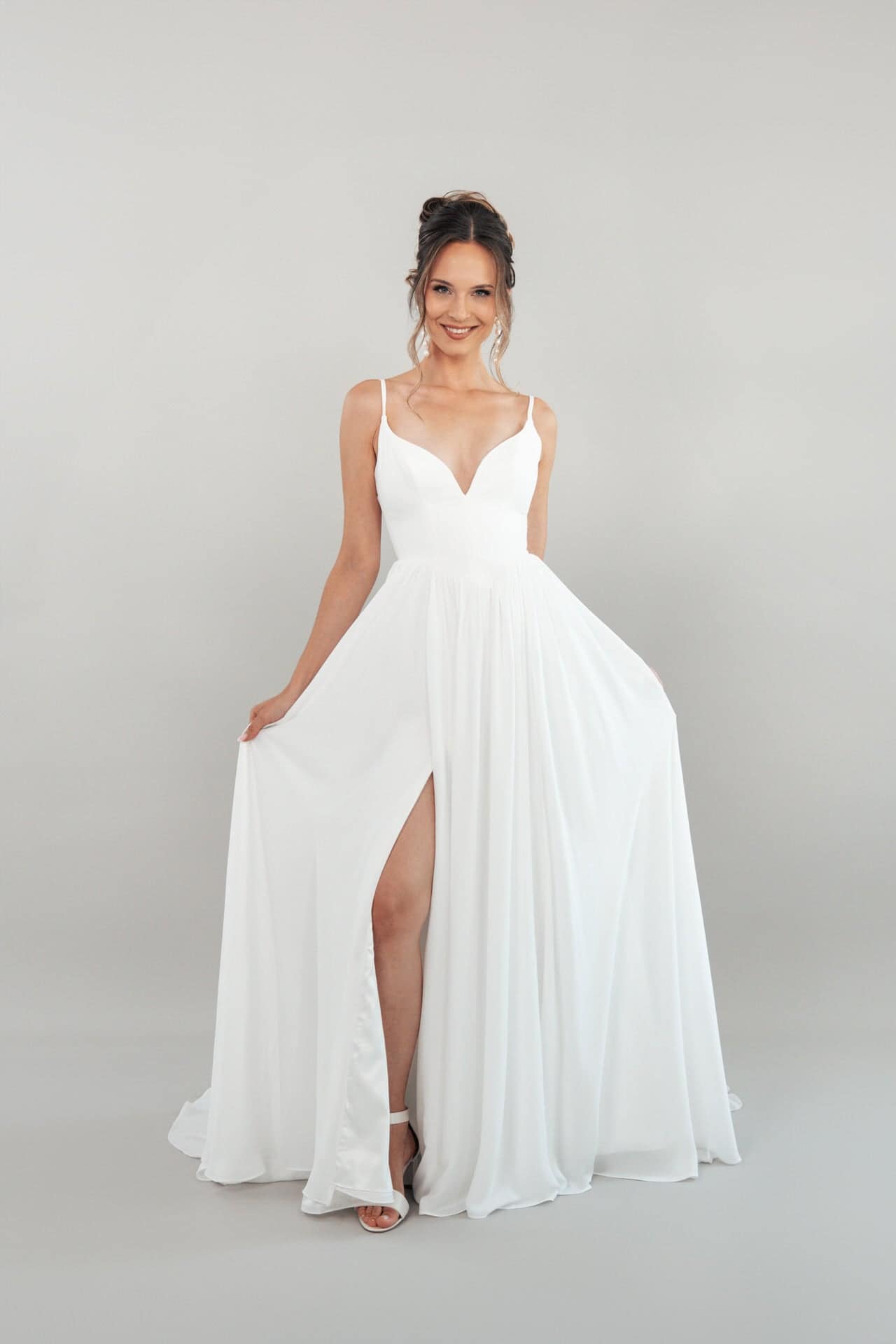 S26-A03_Web_1 Das Modell trägt das S26-A03, ein weißes, ärmelloses Kleid mit tiefem Ausschnitt und hohem Schlitz, und lächelt, während sie die Seiten des Kleides vor einem schlichten hellgrauen Hintergrund hält.