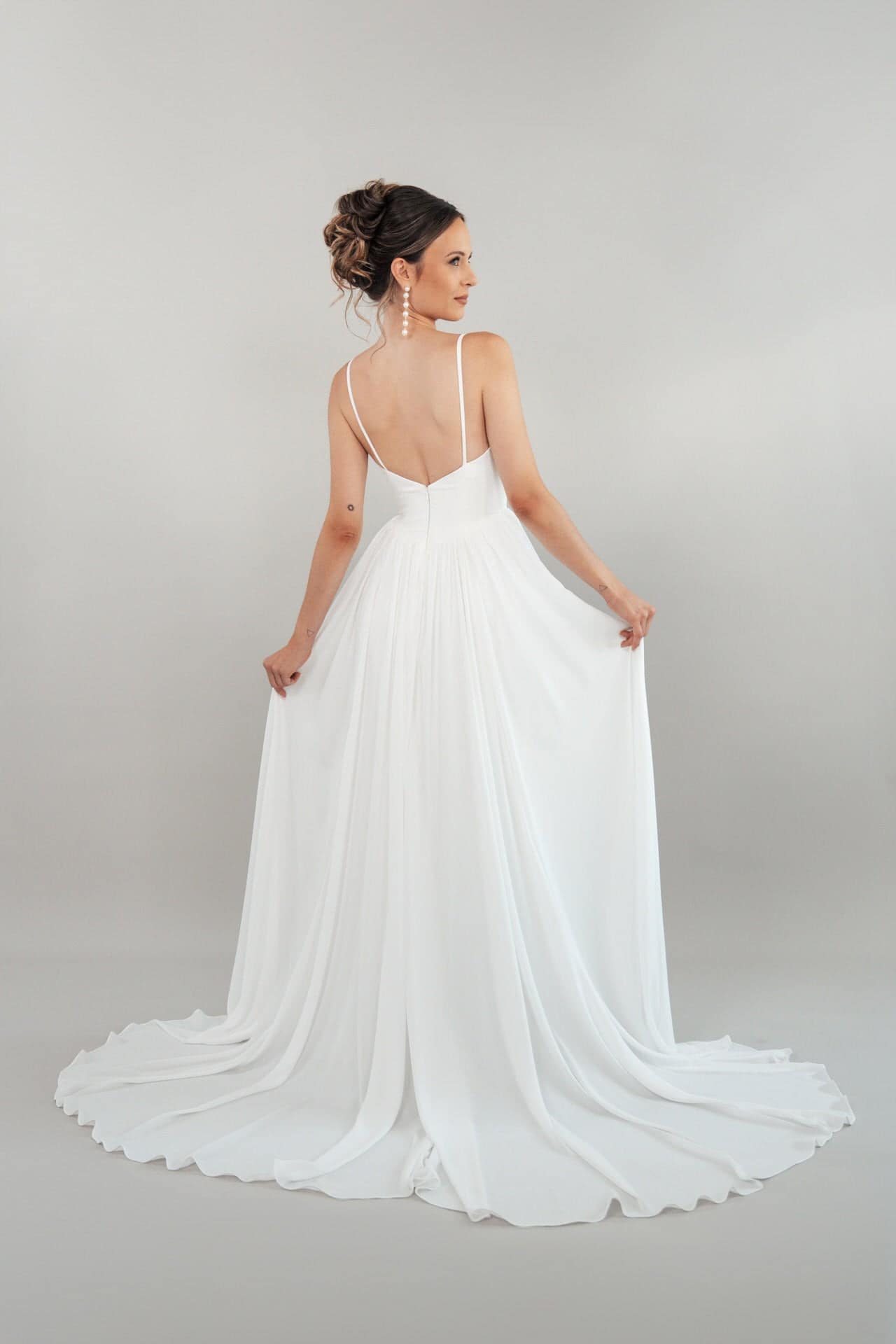 S26-A03_Web_3 Eine Frau steht mit dem Gesicht zur Wand und hält sich an den Seiten ihres Hochzeitskleides S26-A03 fest. Das elegante weiße Kleid hat dünne Träger, einen tiefen V-Ausschnitt und eine lange, fließende Schleppe vor einem schlichten hellen Hintergrund.