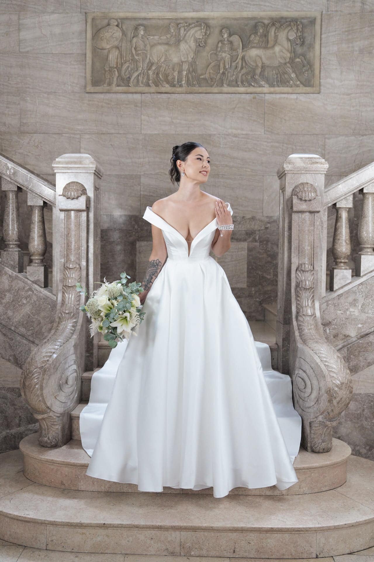 S26-B03_Web_1 Eine Braut in dem schulterfreien weißen Kleid S26-B03 hält einen Blumenstrauß, während sie auf einer großen Steintreppe steht, über der sich ein verziertes Geländer und geschnitzte Kunstwerke befinden.