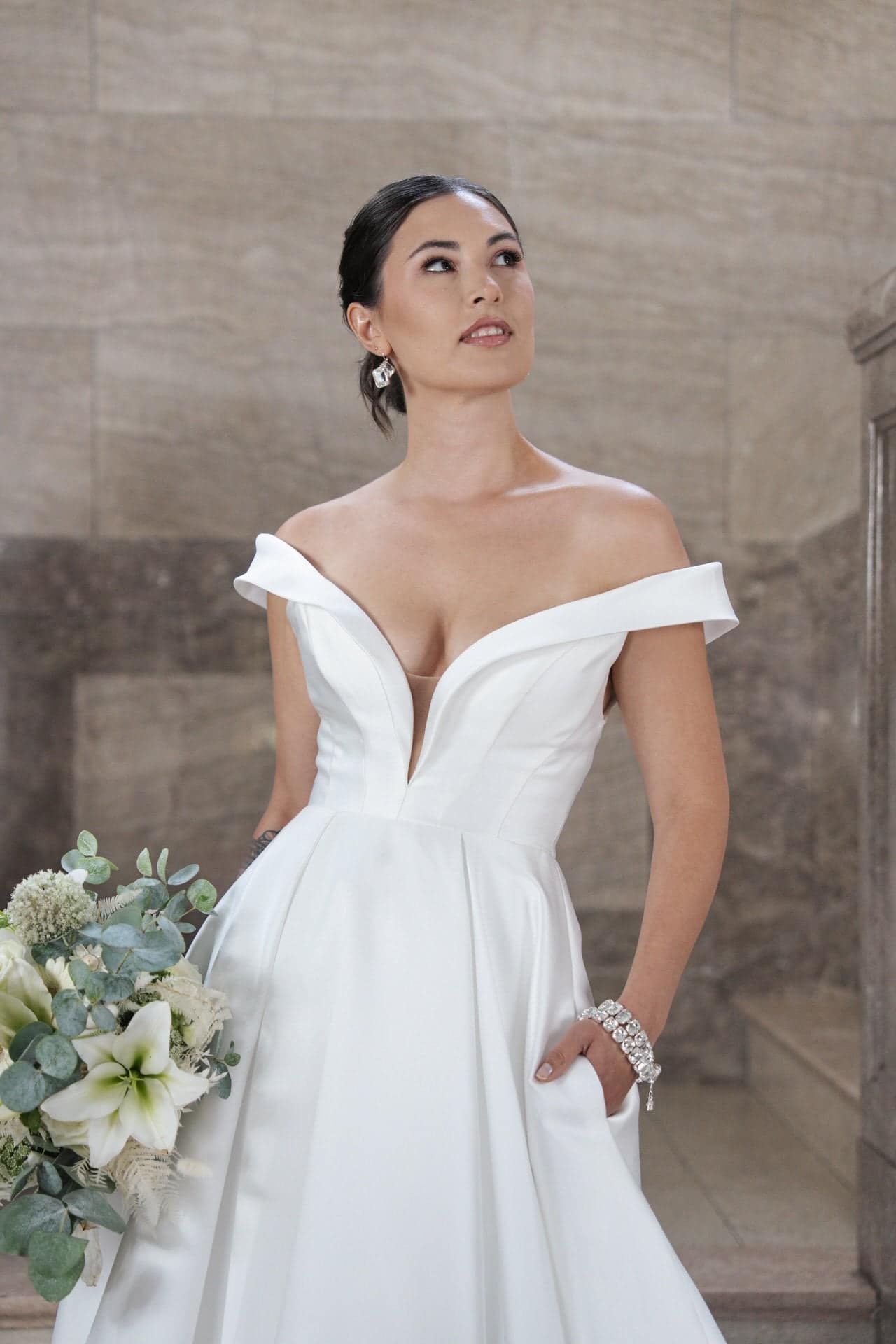 S26-B03_Web_3 Eine Frau trägt das schulterfreie weiße Hochzeitskleid S26-B03 und hält einen Strauß aus weißen Blumen und Grünzeug. Dazu trägt sie Ohrringe und ein Armband. Sie posiert vor einem Hintergrund aus Stein und blickt leicht nach oben.