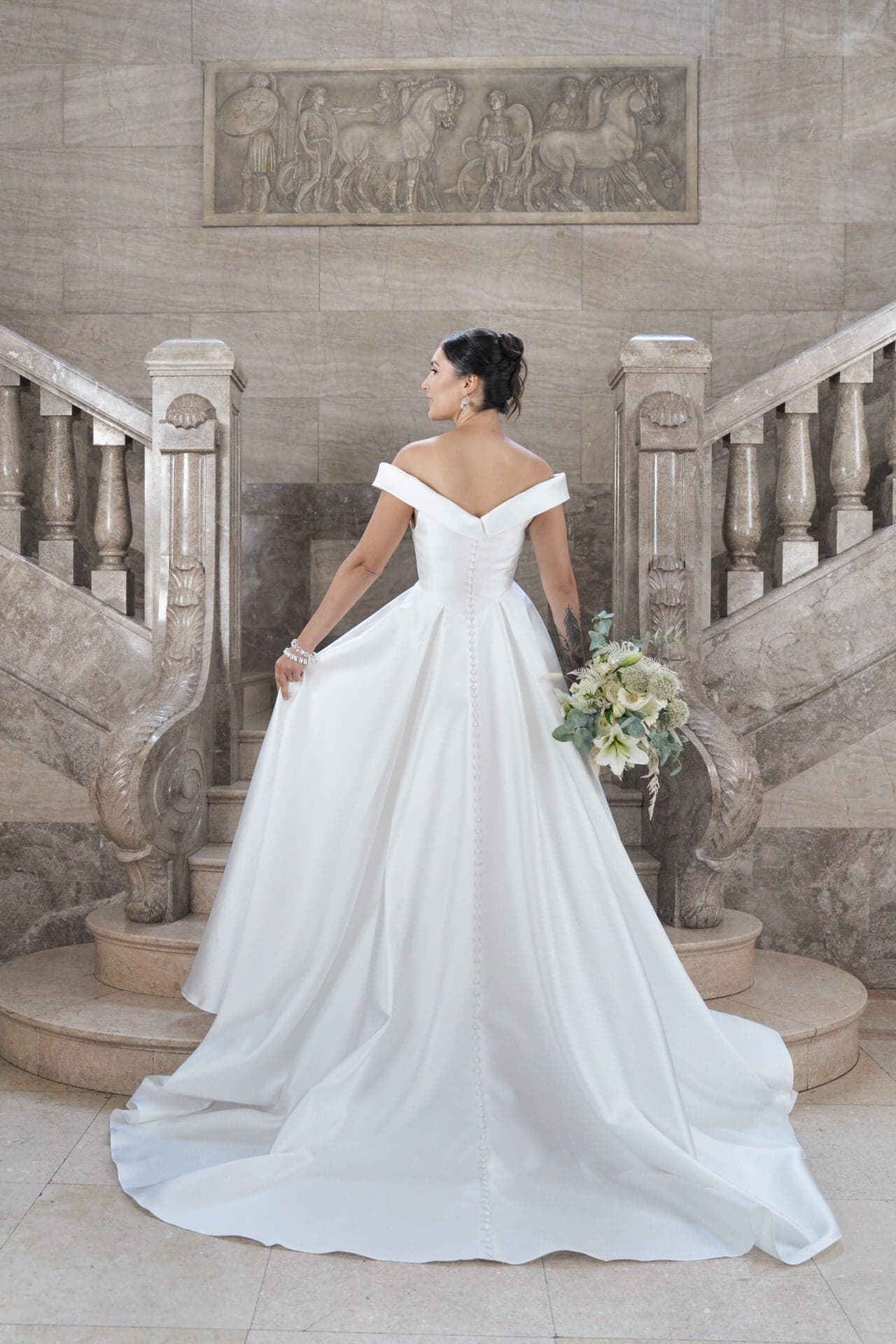 S26-B03_Web_4 Eine Braut in dem schulterfreien weißen Kleid S26-B03 steht auf einer Marmortreppe, hält einen Blumenstrauß in der Hand und blickt über ihre Schulter. Die elegante Treppe und die geschnitzte Wandkunst schaffen einen großartigen, klassischen Rahmen.