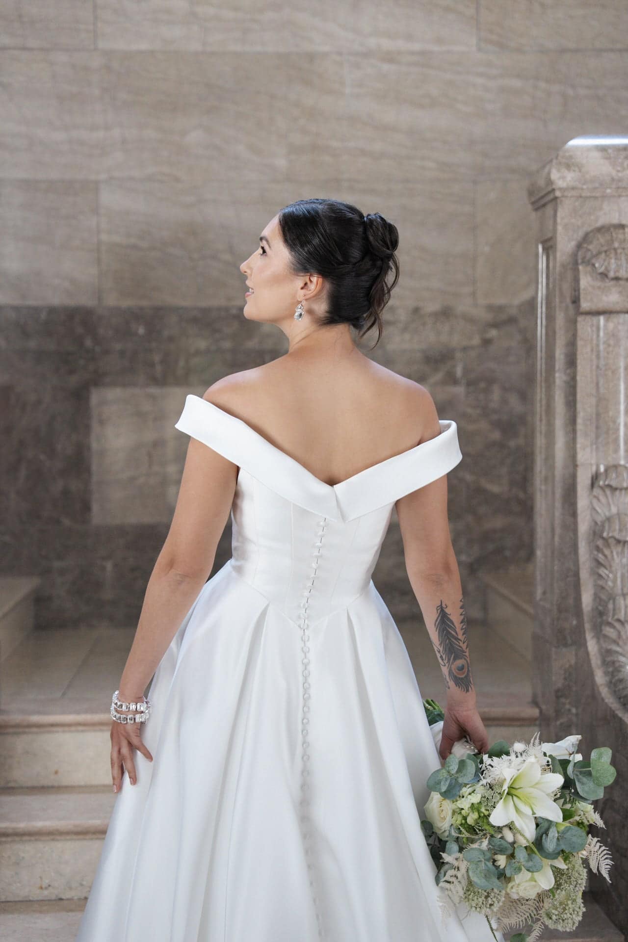 S26-B03_Web_5 In dem schulterfreien weißen Kleid S26-B03 steht eine Braut mit dunklem Dutt und sichtbarem Armtattoo auf einer Treppe und hält einen Strauß mit weißen Blumen und Grünzeug in der Hand, während sie nach oben blickt.