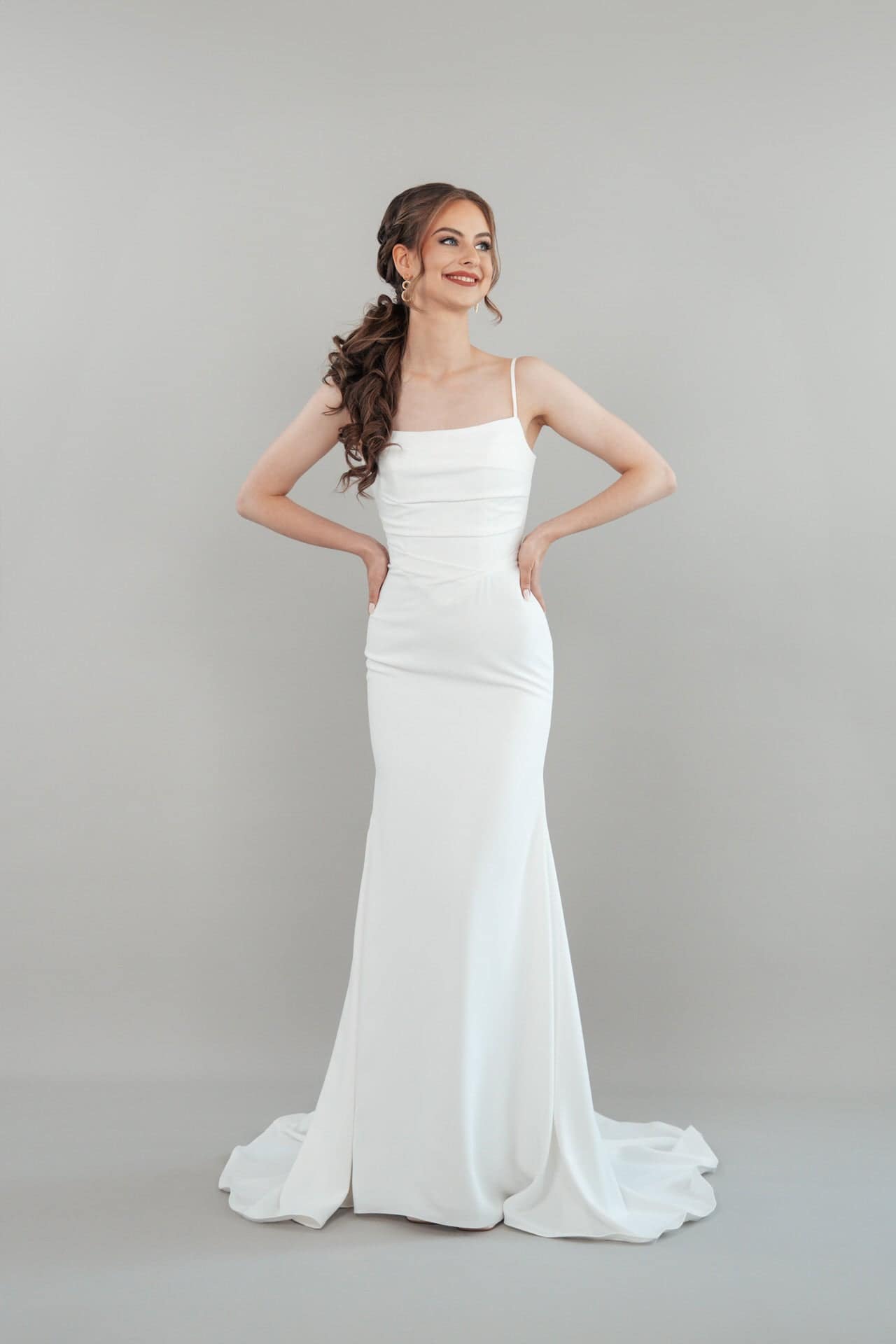 S26-F04_Web_1 Eine Frau trägt das S26-F04, ein weißes, bodenlanges, ärmelloses Hochzeitskleid mit dünnen Trägern, und steht lächelnd mit den Händen in den Hüften vor einem schlichten hellgrauen Hintergrund. Ihr Haar ist in lockeren Wellen gestylt.