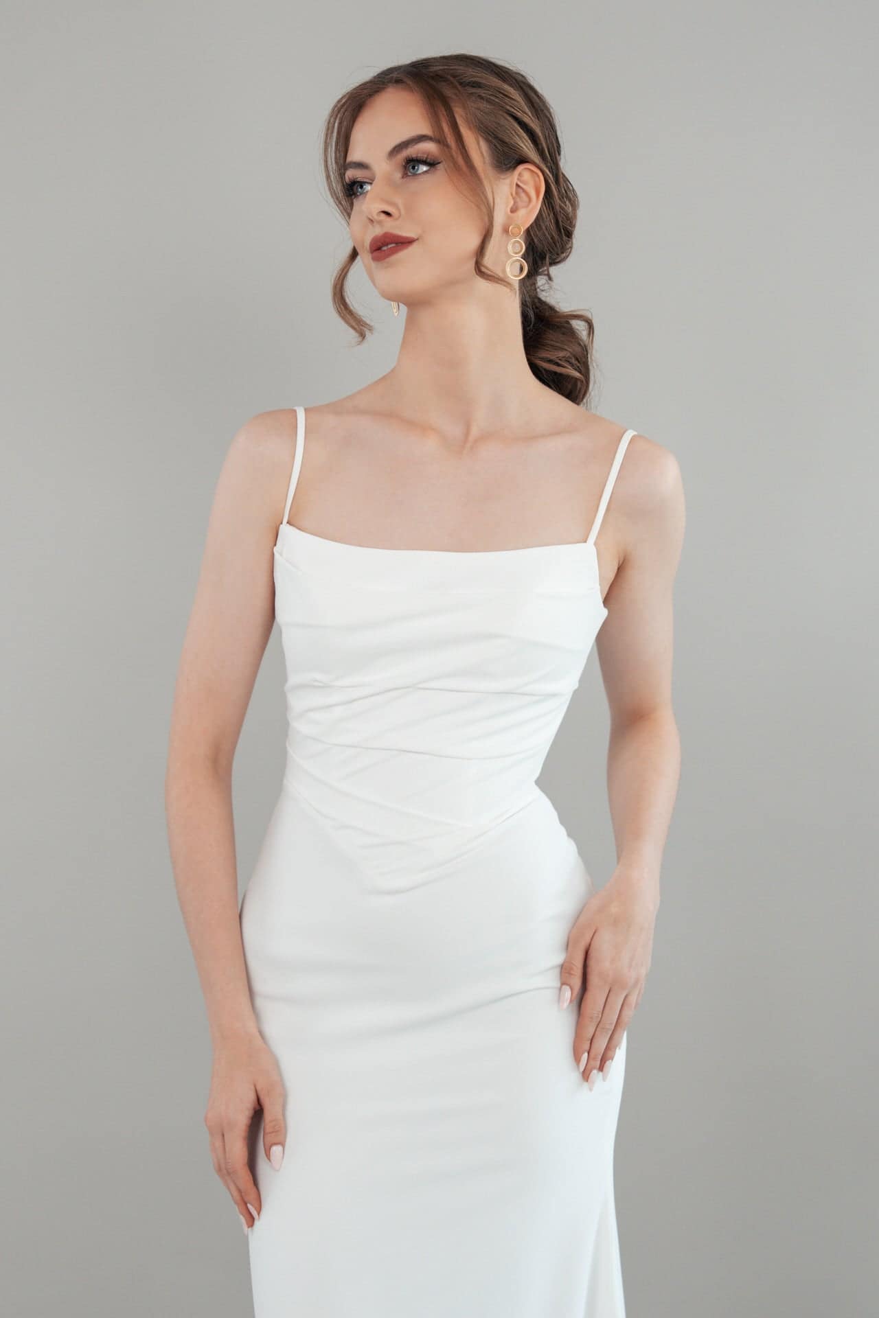 S26-F04_Web_3 Eine Frau mit gewelltem braunem Haar in einer Hochsteckfrisur, die Tropfenohrringe und das ärmellose Kleid S26-F04 mit dünnen Trägern trägt, posiert anmutig vor einem schlichten hellgrauen Hintergrund.