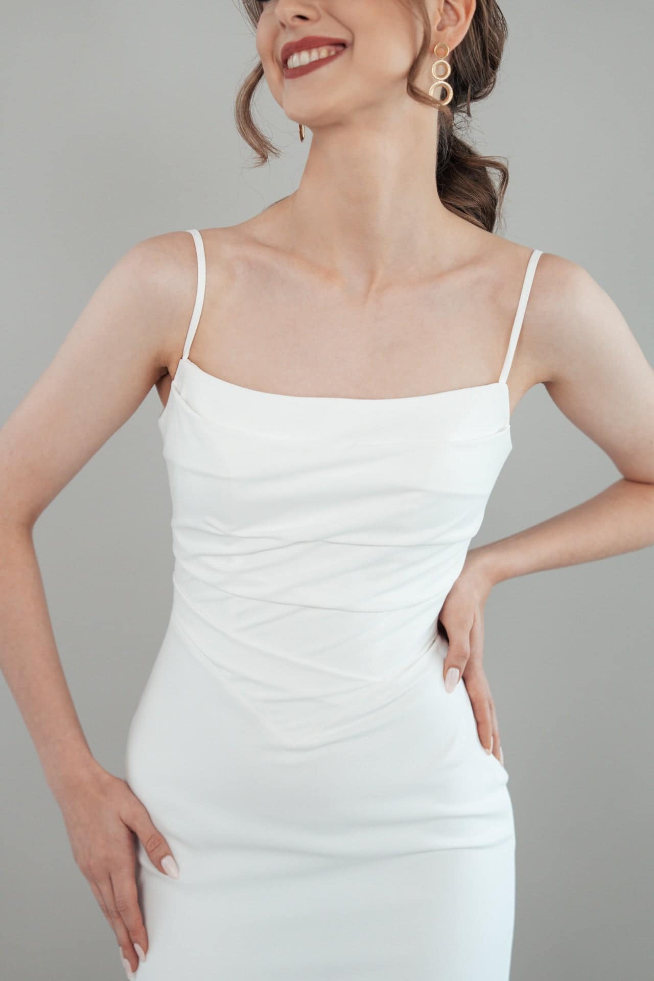 S26-F04_Web_4 Eine Frau in einem weißen Kleid mit dünnen Trägern S26-F04 lächelt mit den Händen in den Hüften. Sie hat gewelltes braunes Haar in einem Pferdeschwanz und trägt große goldene geometrische Ohrringe. Der Hintergrund ist einfarbig hellgrau.