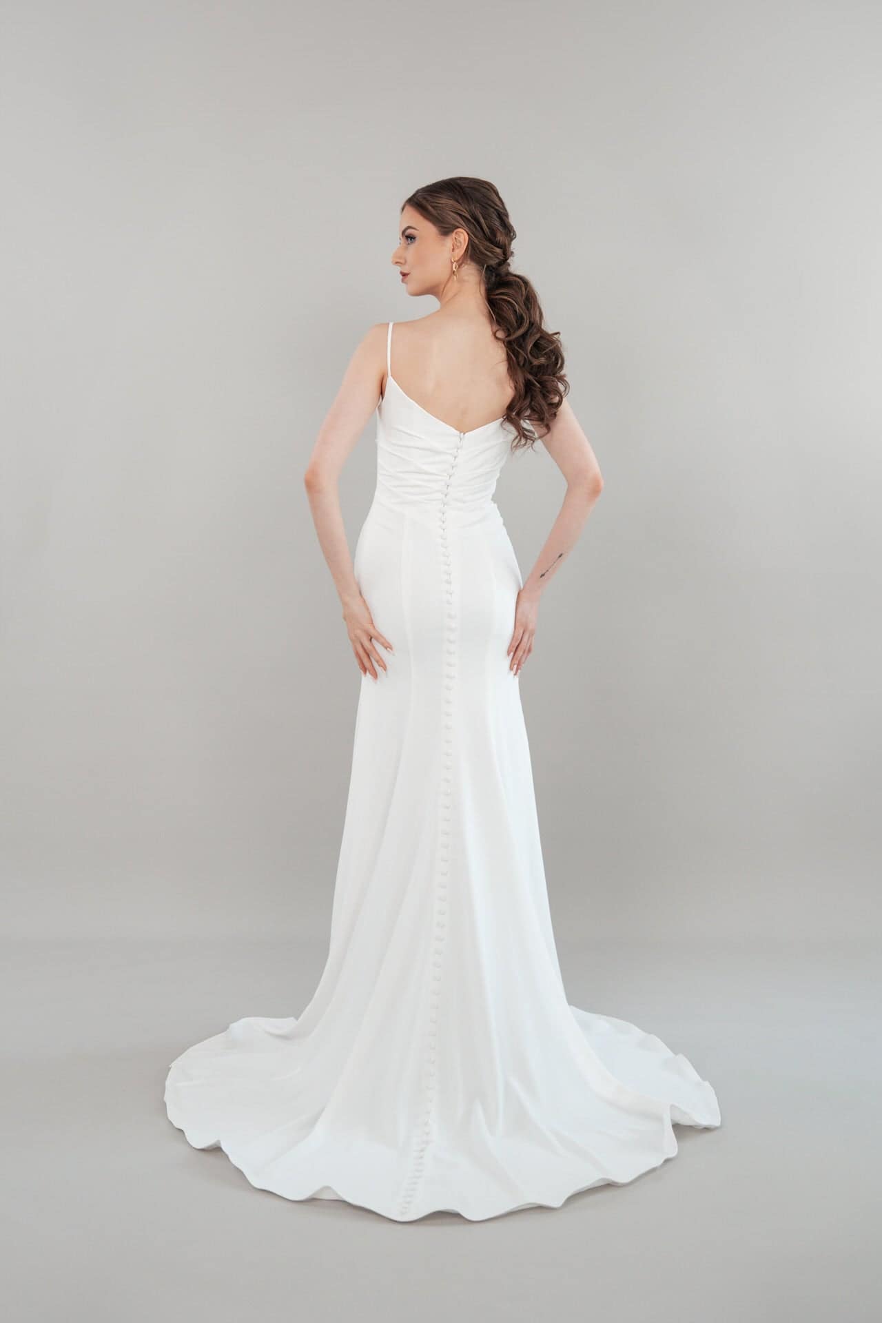 S26-F04_Web_5 Eine Frau mit langem, gewelltem Haar steht vor einem schlichten Hintergrund und präsentiert das S26-F04: ein weißes, bodenlanges, ärmelloses Kleid mit dünnen Trägern, einem taillierten Mieder, einem geknöpften Rücken und einer eleganten Schleppe.