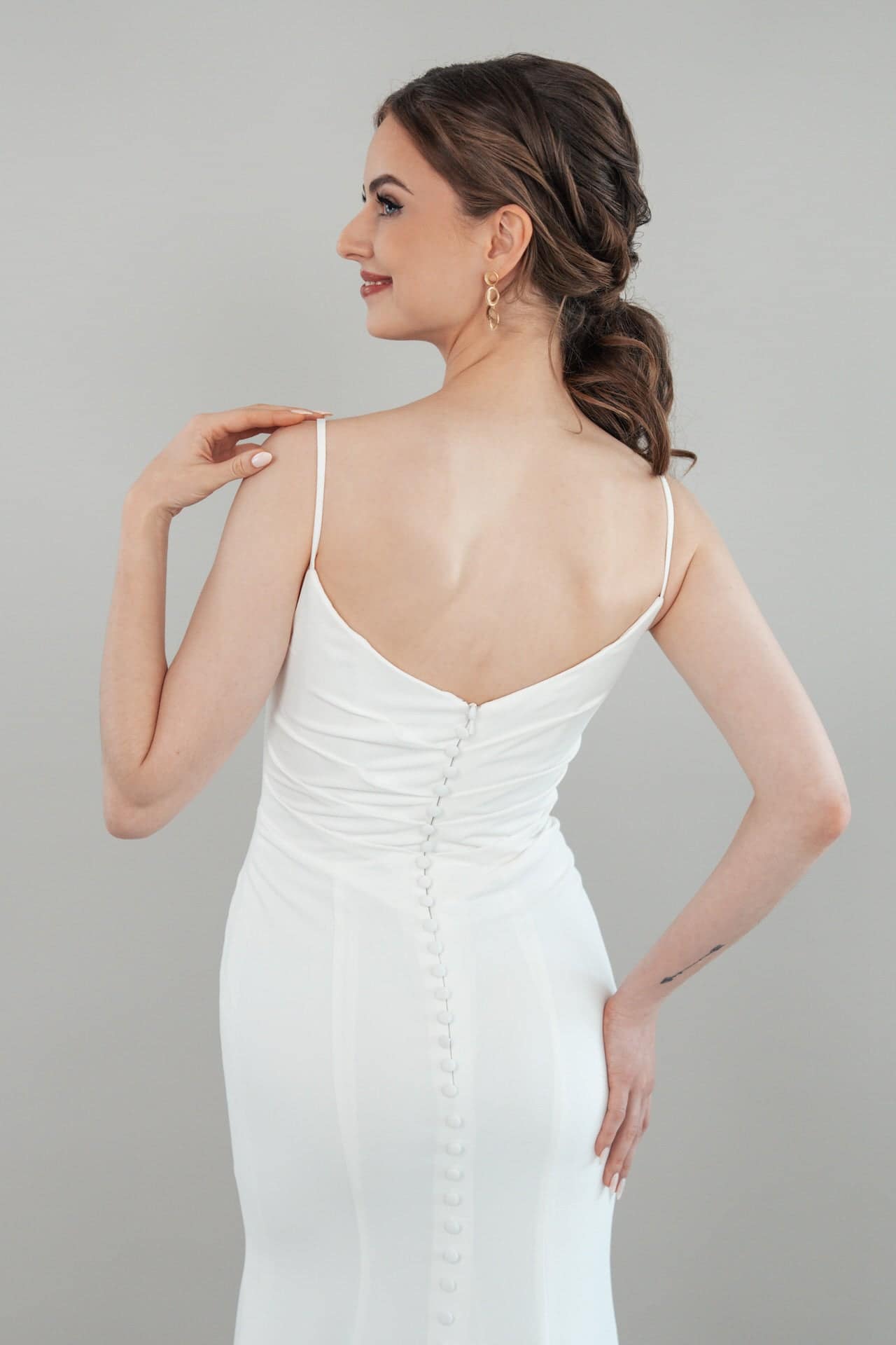 S26-F04_Web_6 Eine Frau, die S26-F04, ein weißes ärmelloses Kleid mit Knopfleiste, trägt, steht zur Seite gedreht und lächelt. Ihr Haar ist in lockeren Wellen nach hinten gestylt, und sie trägt goldene Ohrringe. Der Hintergrund ist einfarbig hellgrau.