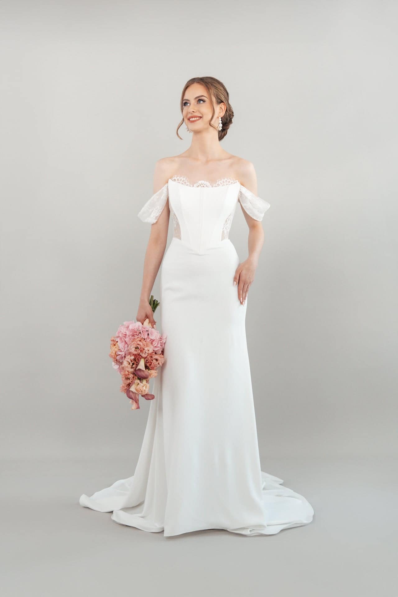 S26-F09_Web_1 Eine Frau, die das schulterfreie weiße Hochzeitskleid S26-F09 trägt, hält einen Strauß rosa Blumen und lächelt vor einem schlichten hellgrauen Hintergrund.