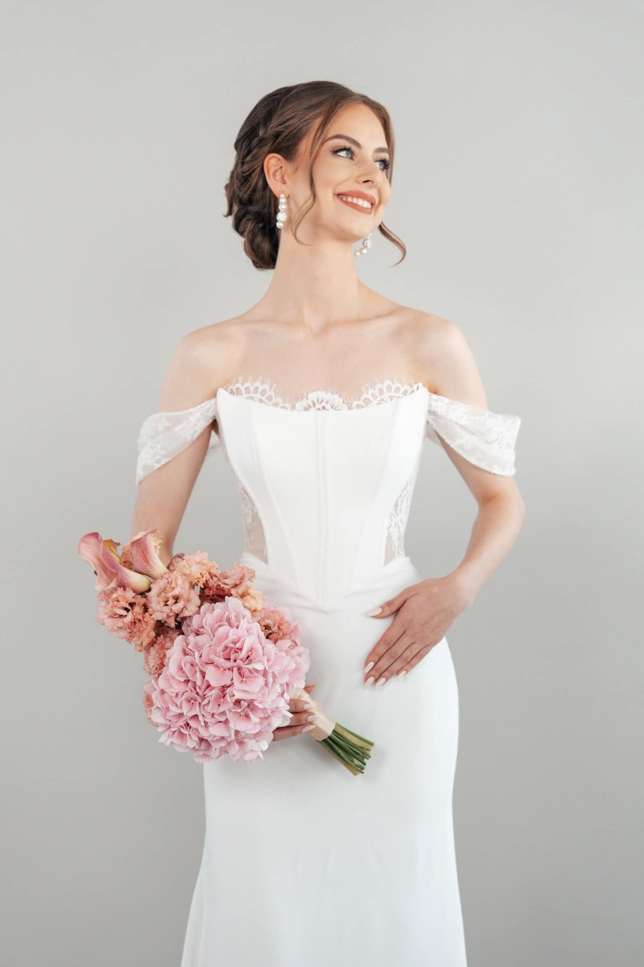 S26-F09_Web_2 Eine lächelnde Braut im schulterfreien weißen Hochzeitskleid S26-F09 hält einen Strauß rosafarbener Blumen und blickt zur Seite vor einem schlichten hellen Hintergrund.
