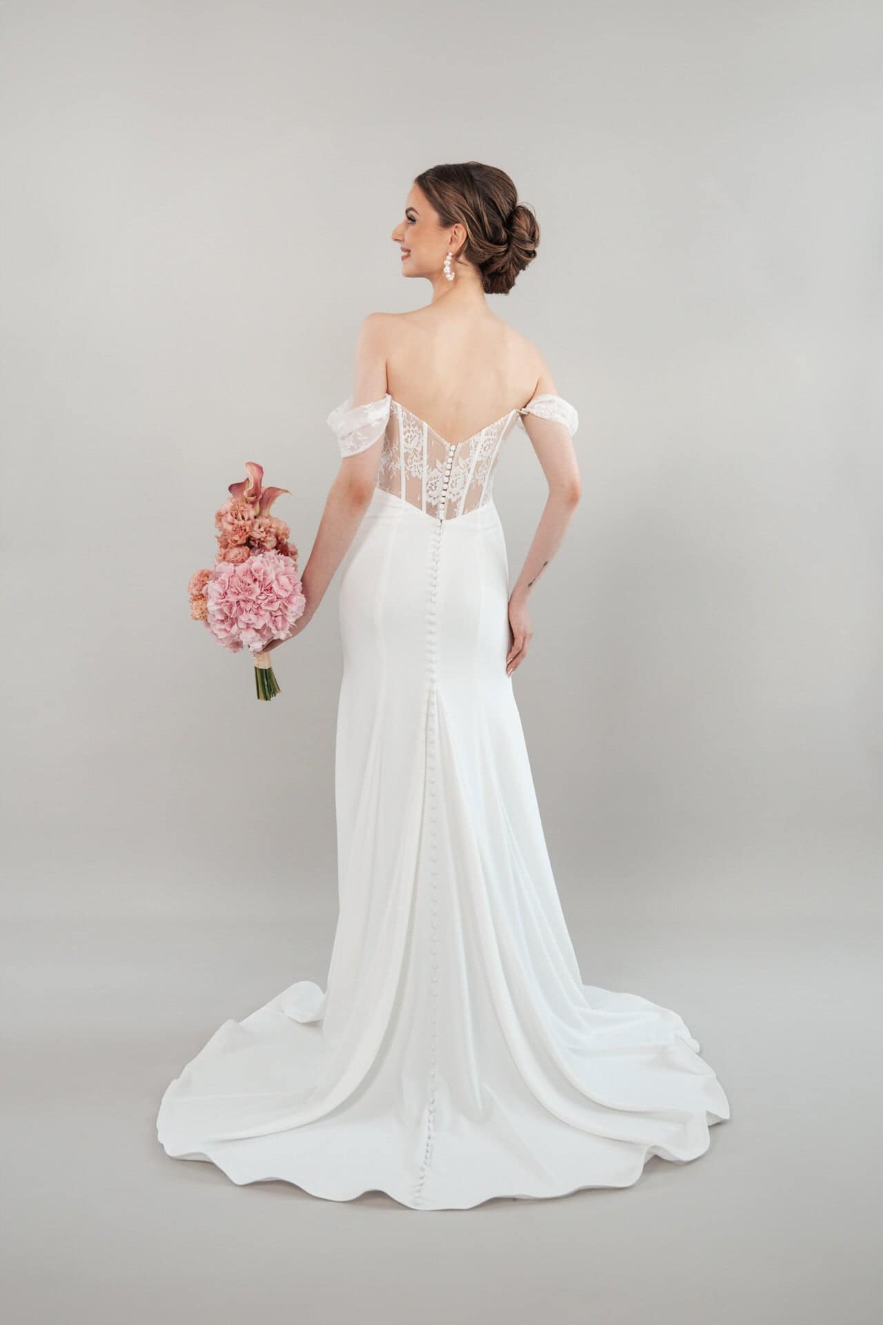 S26-F09_Web_4 Eine Frau steht in S26-F09, einem eleganten, schulterfreien weißen Hochzeitskleid mit Spitzenbesatz im Rücken und Knöpfen, und hält einen Strauß rosa Blumen vor einem hellgrauen Hintergrund.