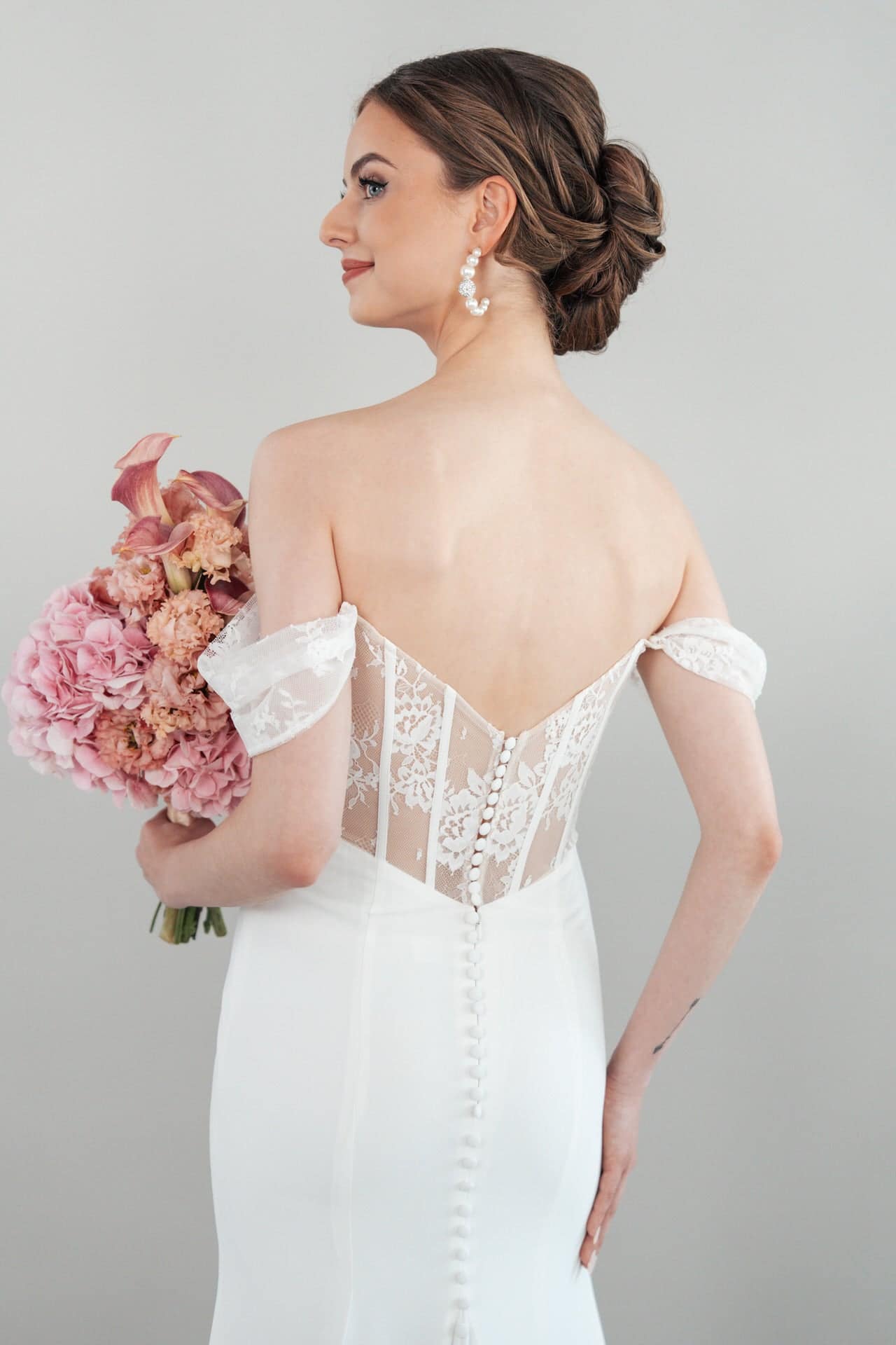 S26-F09_Web_5 Vor einem schlichten Hintergrund steht eine Braut im schulterfreien weißen Brautkleid S26-F09 mit Spitzenverzierung am Rücken, die Haare zu einer eleganten Hochsteckfrisur frisiert, mit Perlenohrringen und einem Strauß rosa Blumen in der Hand.