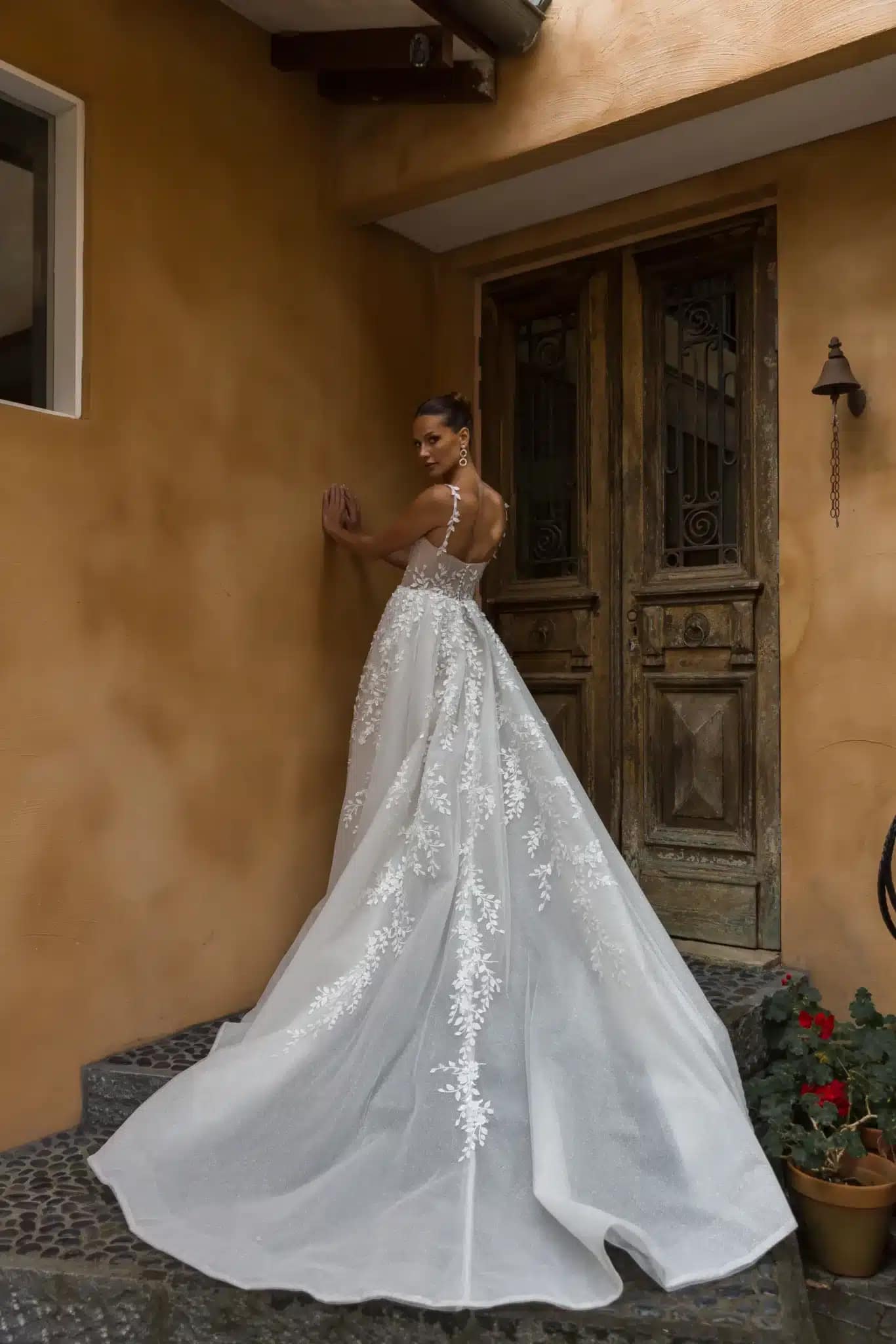 PIA_ML23114_4 Eine Frau in dem Madi Lane Pia Brautkleid steht auf Steinstufen vor alten Holztüren. Die spitzenverzierte Schleppe und die durchsichtigen Träger des Kleides setzen den eleganten Rücken in Szene, während sie die Wand berührt und zeitlosen Brautcharme versprüht.