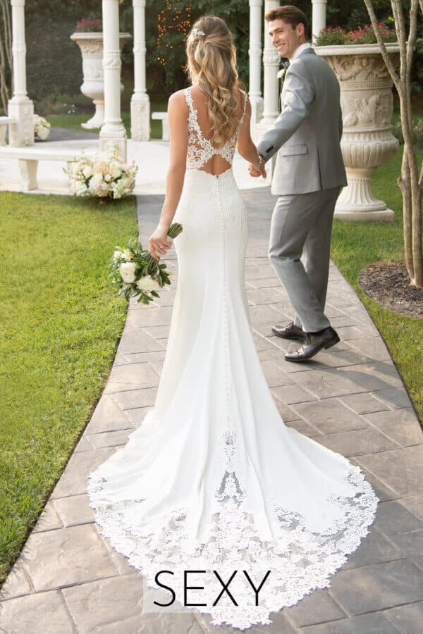 Eine Braut in einem taillierten, rückenfreien Hochzeitskleid hält einen Blumenstrauß und geht Hand in Hand mit einem Bräutigam in einem grauen Anzug einen Gartenweg entlang. Das Wort SEXY erscheint am unteren Rand des Bildes.