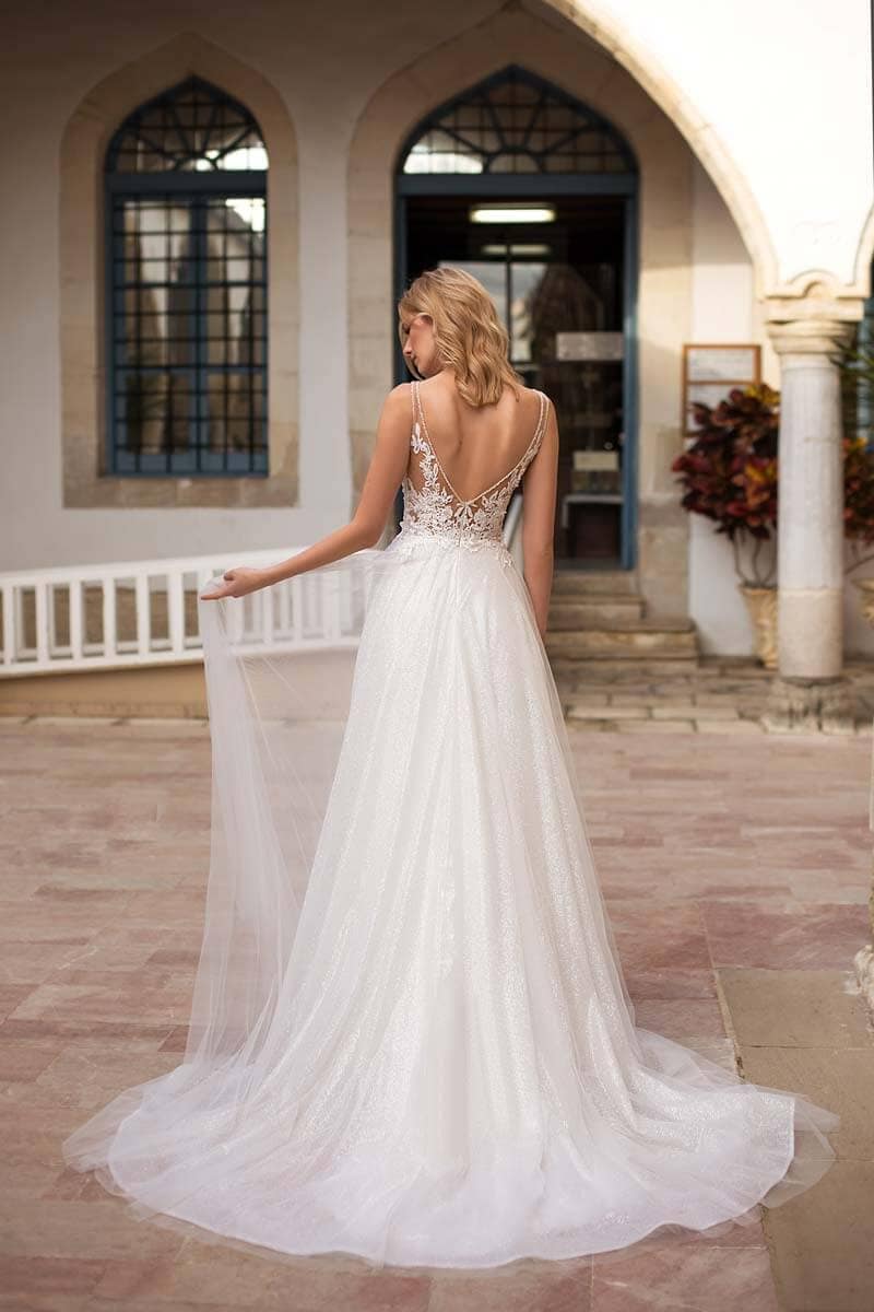 Eine Braut in einem ärmellosen weißen Hochzeitskleid mit tiefem V-Rücken und Spitzendetails steht im Freien und hält einen durchsichtigen Schleier. Im Hintergrund sind Bogenfenster und ein Innenhof zu sehen.