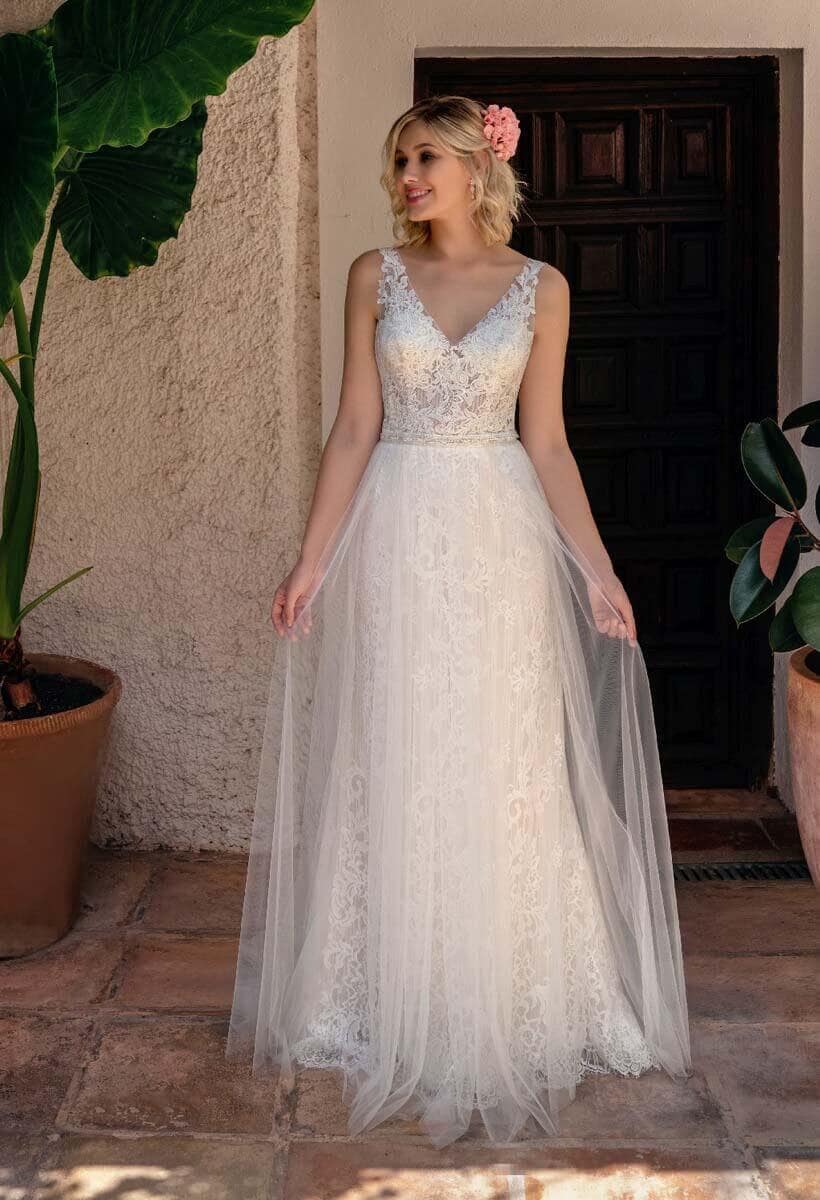 Eine Frau in einem ärmellosen, spitzenbesetzten weißen Hochzeitskleid steht auf einem terrakottafarbenen Fliesenboden und versprüht den Charme von Amelie. Sie hält ihr Kleid, eine rosa Blume im Haar, neben Topfpflanzen an einer strukturierten Wand.