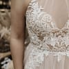 Nahaufnahme einer Person, die das Bridentity Bliss Brautkleid trägt, ein ärmelloses, durchsichtiges Hochzeitskleid mit aufwändiger weißer, floraler Spitzenstickerei auf dem Mieder.