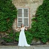 Eine Frau trägt das elegante Bridonista V24-22 Brautkleid und posiert mit leicht erhobenen Armen vor einer alten, efeubewachsenen Steinmauer neben einem geschlossenen, verwitterten Holzfenster.