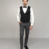 Ein Mann mit kurzen dunklen Haaren in formeller Kleidung, der Masculini 3397-08 trägt, steht vor einem grauen Hintergrund. Neben ihm bilden elegante Brautkleider und Hochzeitskleider den idealen Rahmen für ein stilvolles Hochzeitsensemble.