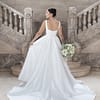 Die Braut trägt das Hochzeitskleid S26-B02 und steht mit dem Rücken zur Kamera am Fuße einer großen Steintreppe, in der Hand einen Strauß weißer und grüner Blumen. Das Kleid hat eine lange Schleppe und ist mit eleganten Knöpfen verziert.