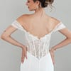 Eine Frau mit einer eleganten Hochsteckfrisur modelliert das S26-F07, ein schulterfreies, weißes Hochzeitskleid mit Spitzenrücken. Sie posiert mit dem Rücken zur Kamera und den Händen in den Hüften vor einem schlichten Hintergrund.