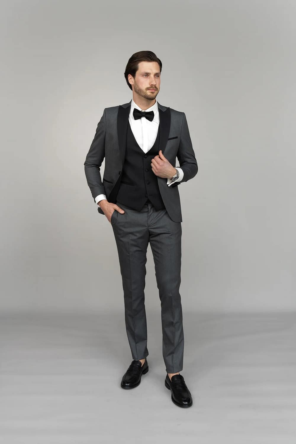 Ein Mann steht vor einem schlichten grauen Hintergrund und trägt den Anzug Masculini 3397-08, eine stilvolle Wahl, die perfekt zu jedem Brautkleid oder Hochzeitskleid passt, mit einer Hand in der Tasche und einem Blick zur Seite.