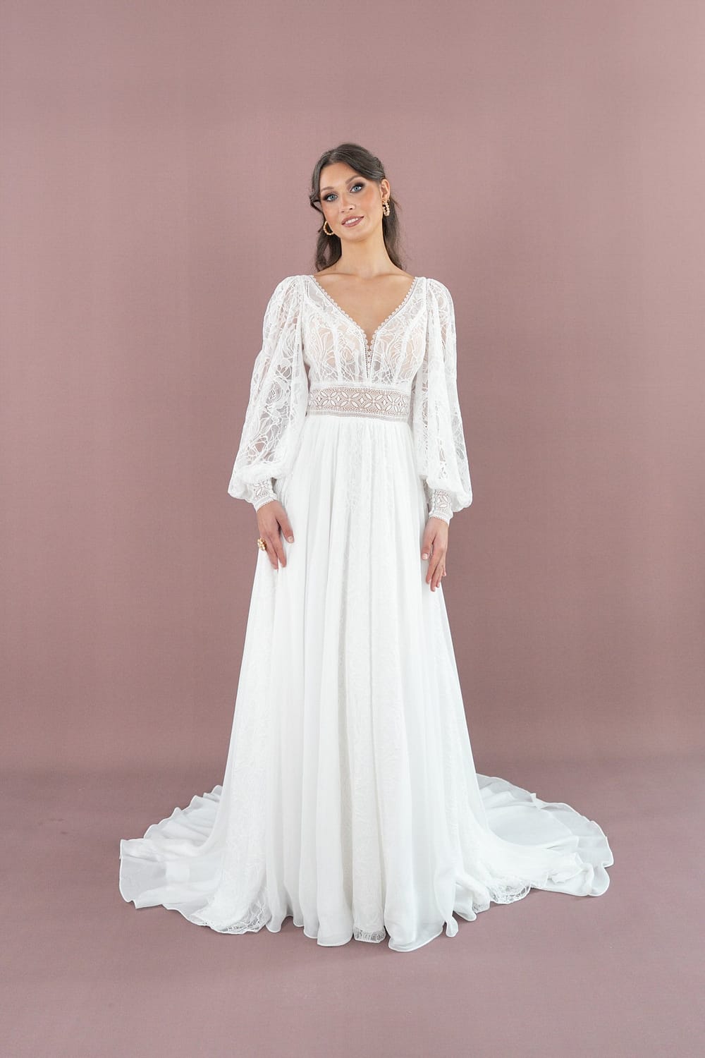 Eine Frau steht vor einem mauvefarbenen Hintergrund und trägt das Bridonista V26-A10, ein langes weißes Brautkleid mit durchsichtigen Spitzenärmeln, tiefem V-Ausschnitt, tailliertem Spitzenmieder und fließendem Rock mit Schleppe - eine elegante, moderne Brautmodenwahl.