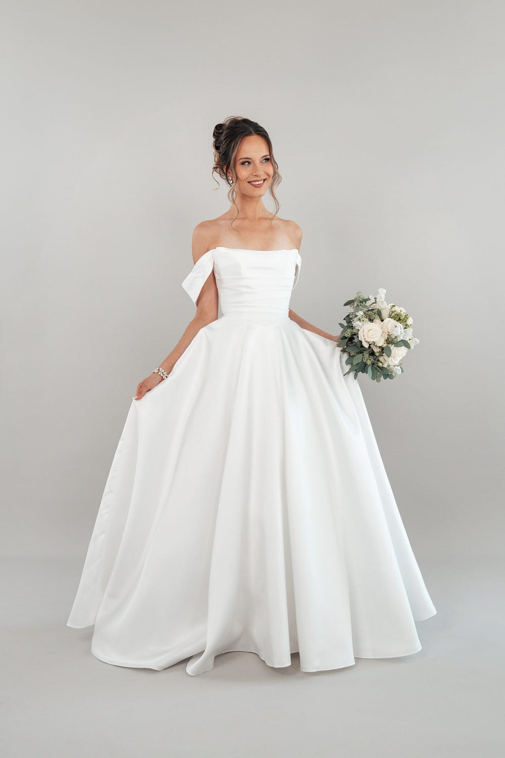 Eine lächelnde Frau trägt das schulterfreie weiße Brautkleid S26-B01 und hält einen Strauß weißer und grüner Blumen vor einem schlichten hellgrauen Hintergrund.
