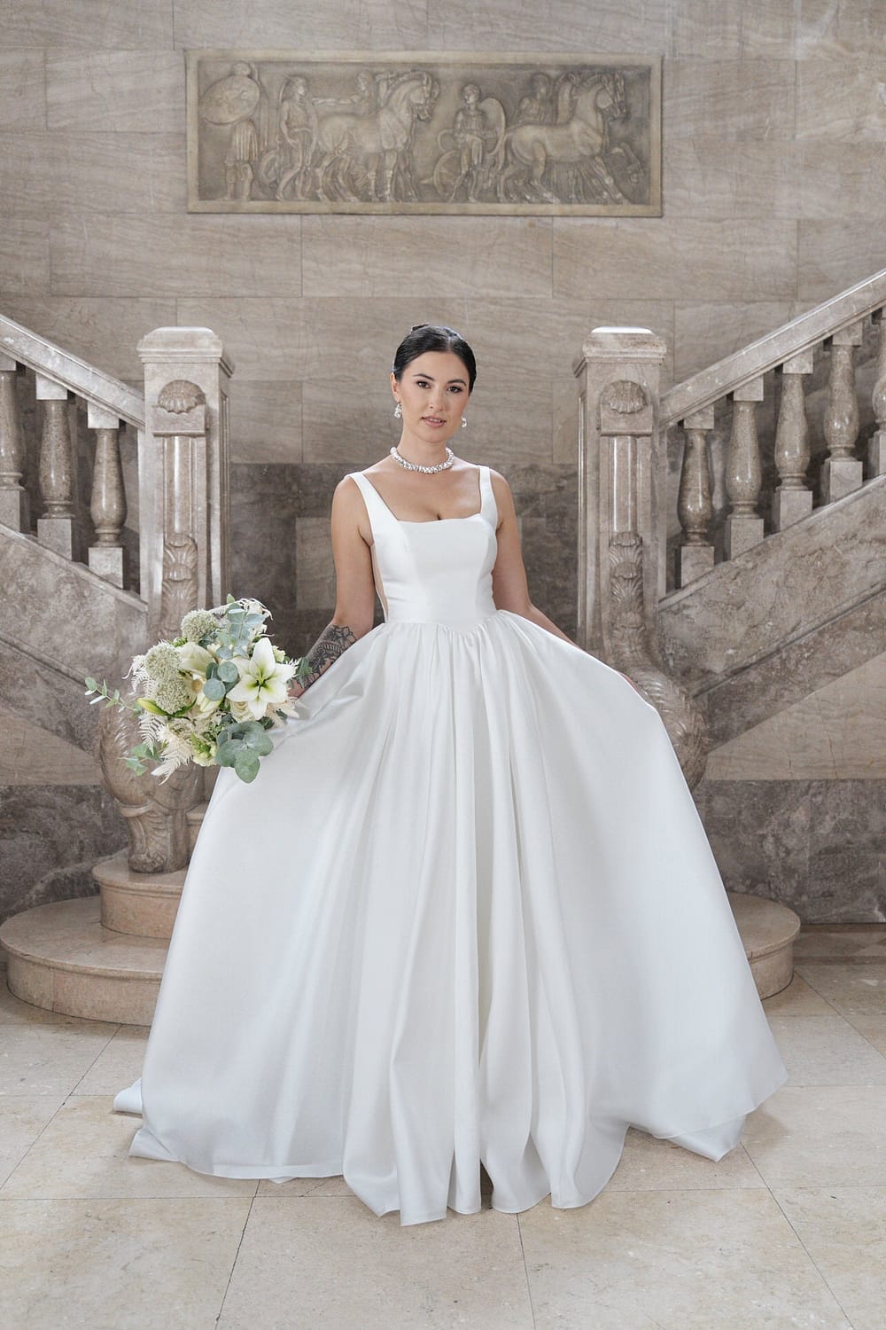 Eine Frau, die das weiße Hochzeitskleid S26-B02 trägt, hält einen Blumenstrauß am Fuße einer großen Marmortreppe mit kunstvollem Geländer und einem geschnitzten Wandrelief hinter ihr.