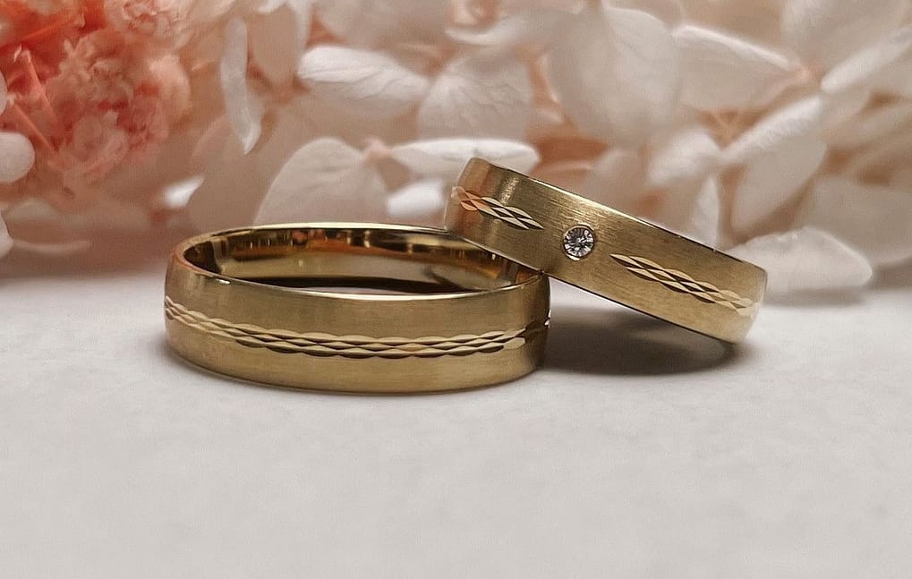 Zwei goldene Eheringe mit eingravierten Mustern liegen auf einer weißen Fläche; ein Ring ist mit einem kleinen Diamanten besetzt, während zartrosa und weiße Blumen - die Inspiration für das perfekte Brautkleid - sanft im Hintergrund verschwimmen.
