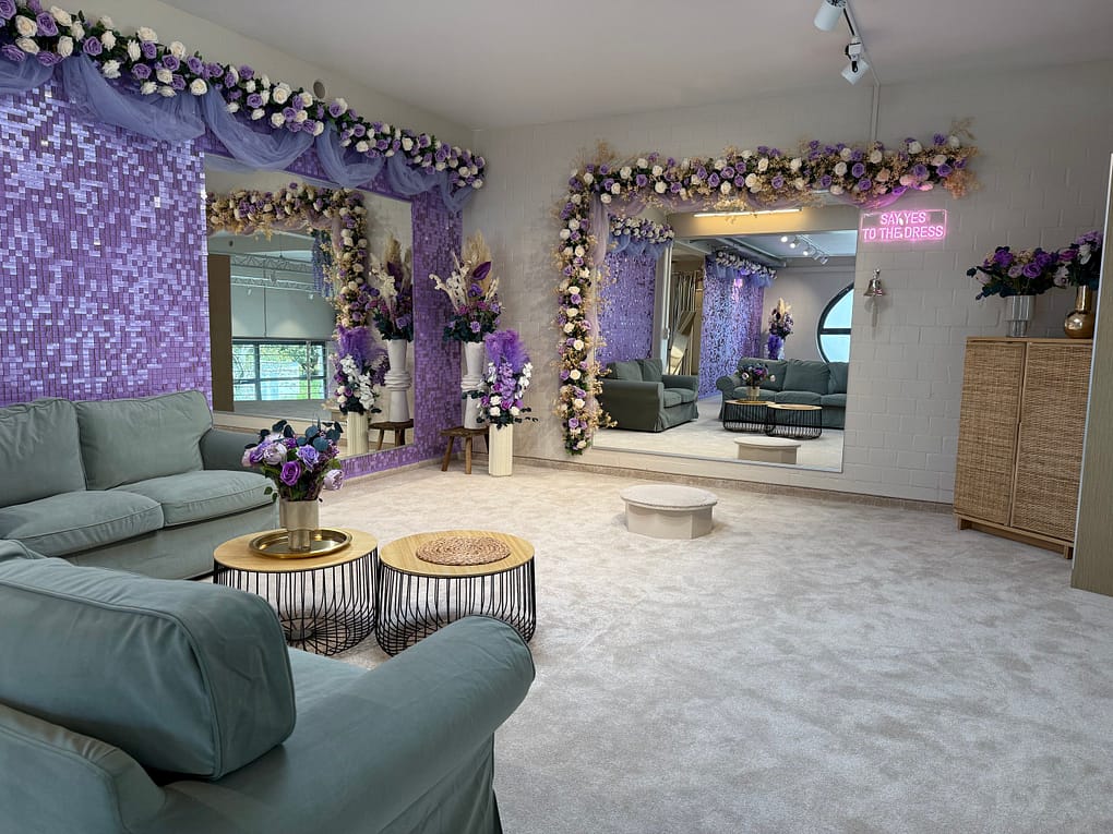 Die stilvolle Lounge im hochzeitsrausch Brautmoden Berlin Schönefeld ist mit hellgrünen Sofas, goldenen und schwarzen Couchtischen und großen, von Blumen eingerahmten Spiegeln ausgestattet. Lila Mosaikfliesen, Blumengirlanden und ein neonfarbenes "SAY YES TO THE DRESS"-Schild vervollständigen das Anprobezimmer.
