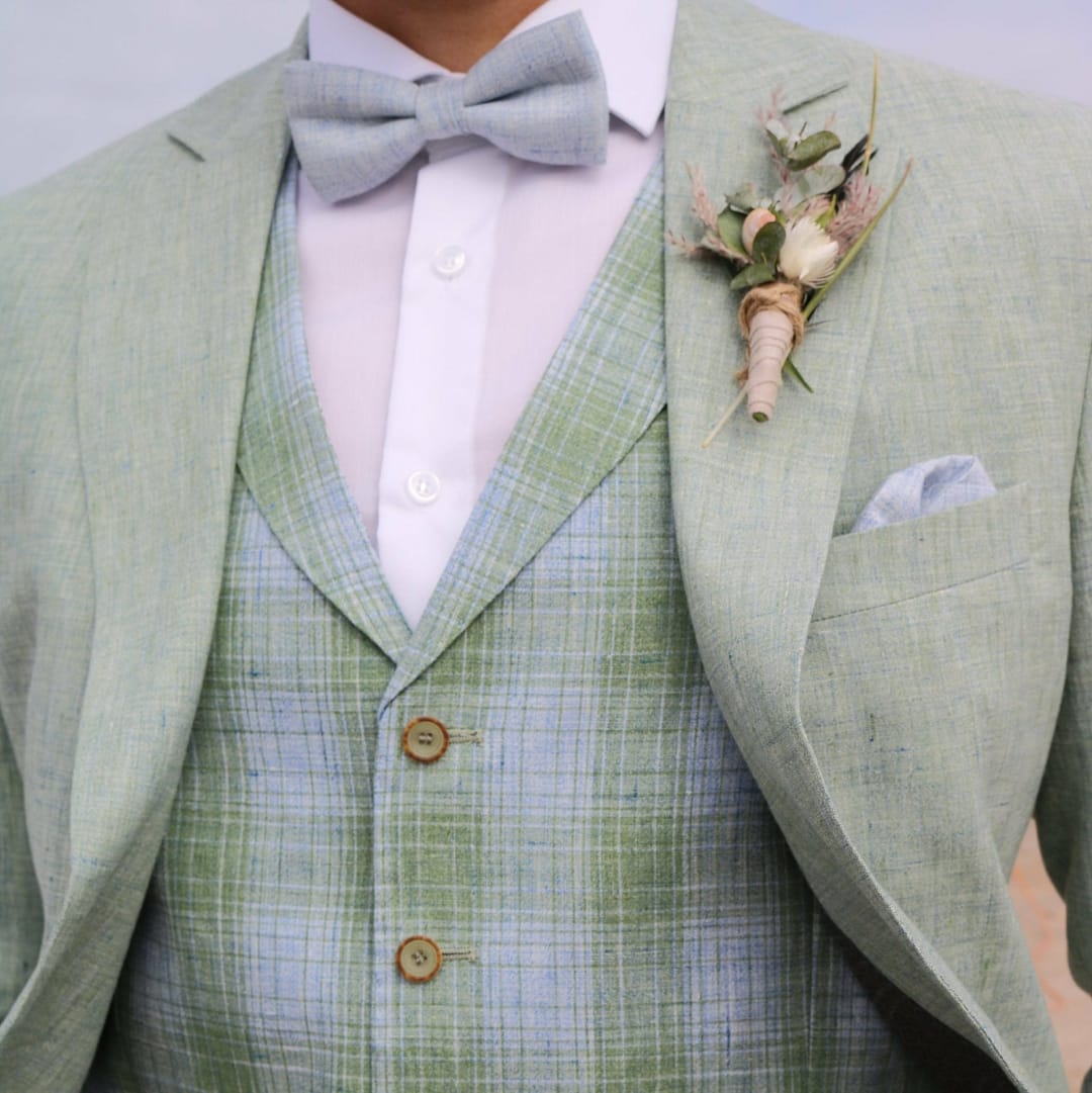 Ein Mann trägt die Atelier Torino Vintage Wedding Schleife / Tuch 821779/30 - einen hellgrünen Anzug mit passender Fliege, grün karierter Weste, Boutonniere und hellblauem Einstecktuch - als einzigartige Hochzeitskleid-Inspiration; nur sein Oberkörper ist sichtbar.