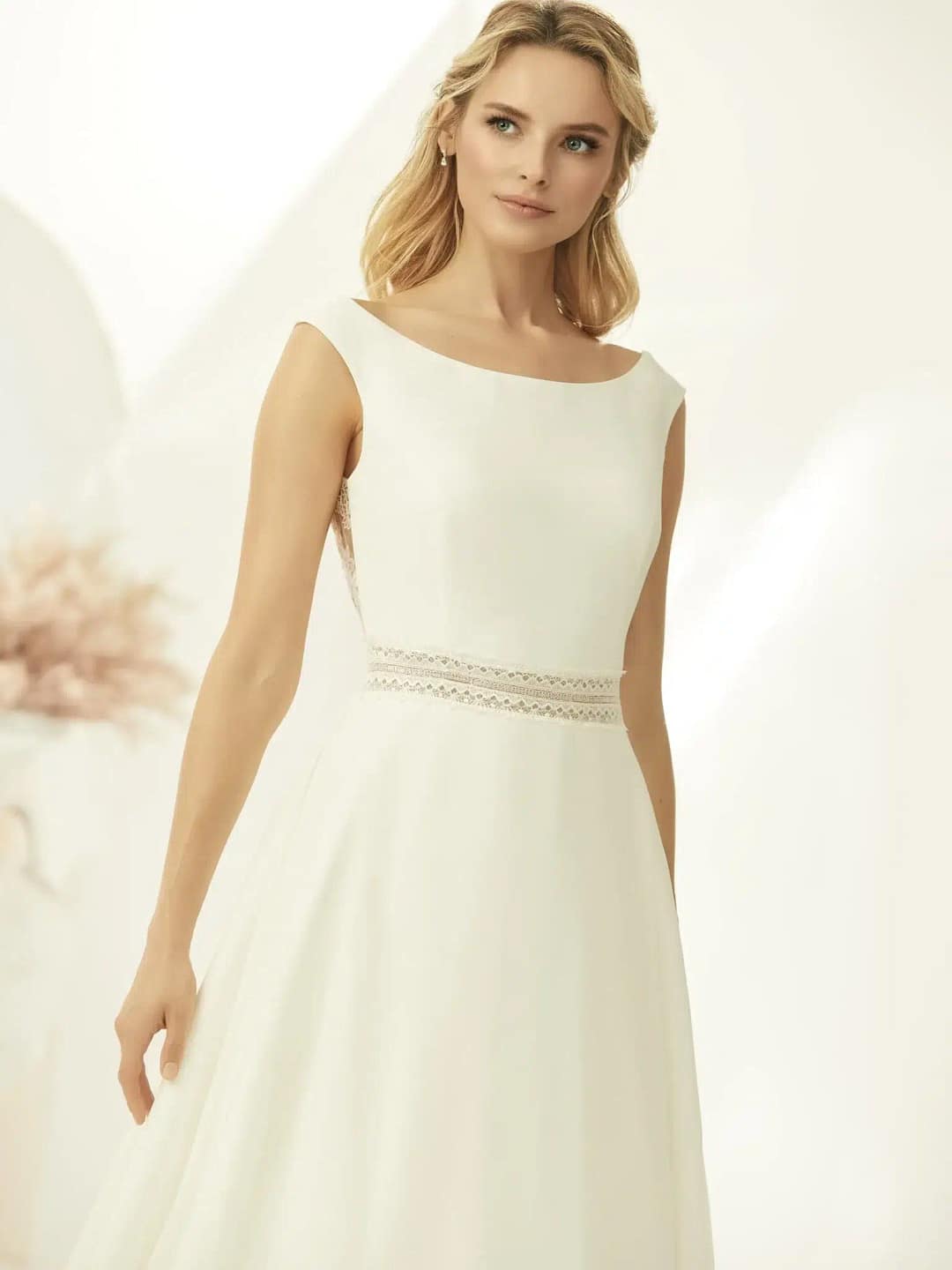 Bianco Evento Parma Brautkleid Hochzeitskleid-2 Eine Frau im Bianco Evento Parma Brautkleid, einem elfenbeinfarbenen ärmellosen Kleid mit Bateau-Ausschnitt und Spitzendetail an der Taille, steht vor einem sanft beleuchteten neutralen Hintergrund und verkörpert zeitlose Eleganz.