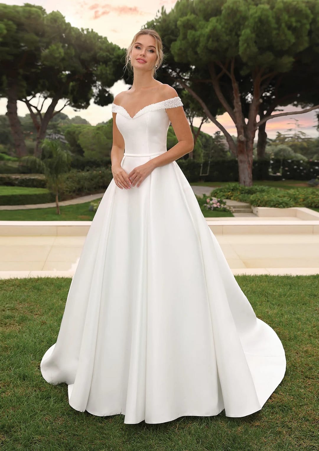 Ladybird-LYNNIE-B Eine Frau steht im Freien in einem Garten und trägt das elegante Ladybird Lynnie Hochzeitskleid mit tailliertem Mieder und vollem Rock. Bäume und Grünpflanzen bilden eine romantische Kulisse, die ihren zeitlosen Brautstil unterstreicht.