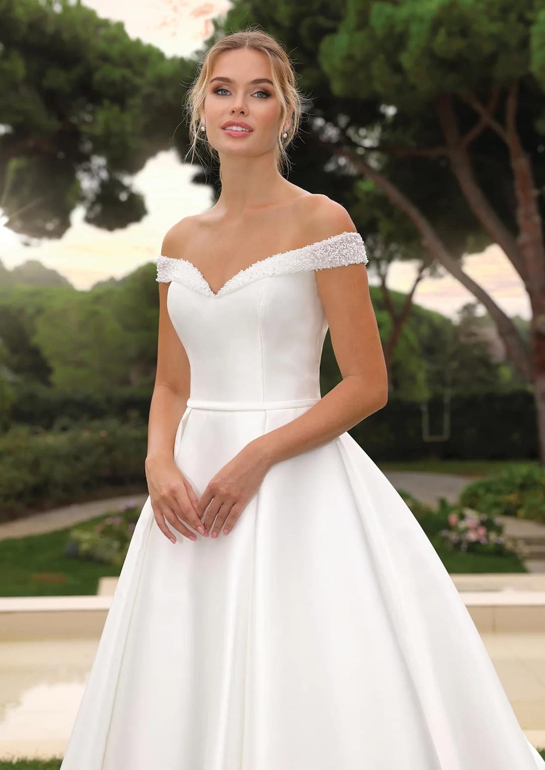 Ladybird-LYNNIE-D Eine Frau in einem schulterfreien Ladybird Lynnie Hochzeitskleid steht im Freien inmitten von Grün und Bäumen, das Haar zu einer Hochsteckfrisur frisiert und mit einem sanften Lächeln im Gesicht, das vom natürlichen Licht sanft beleuchtet wird.