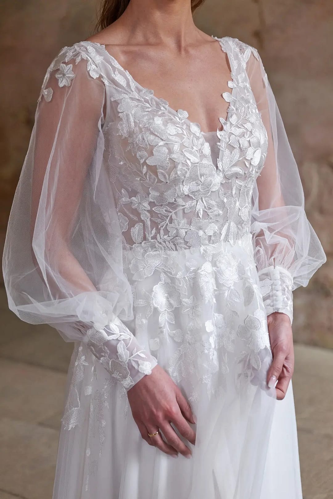 BridonistaBridalV24-24_LQ_4 Eine Frau trägt das Bridonista V24-24 Brautkleid, ein weißes Hochzeitskleid mit durchsichtigen langen Puffärmeln und detaillierten floralen Spitzenapplikationen an Mieder und Ärmeln, vor einem neutralen Hintergrund.