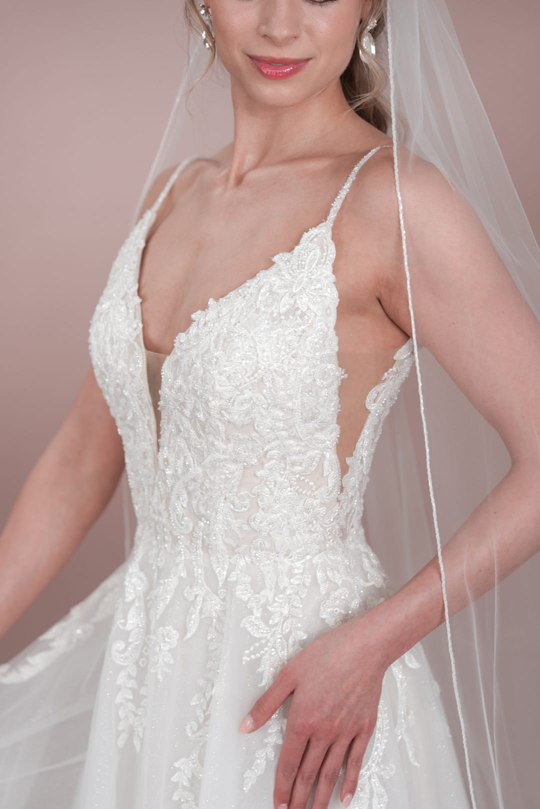 C24-19(CL)_HQ (4) Eine Frau trägt das Bridonista C24-19 Hochzeitskleid, ein weißes Spitzenkleid mit dünnen Trägern, tiefem Ausschnitt und aufwändiger Stickerei, gestylt mit einem Schleier, hellrosa Lippenstift und Perlenohrringen.