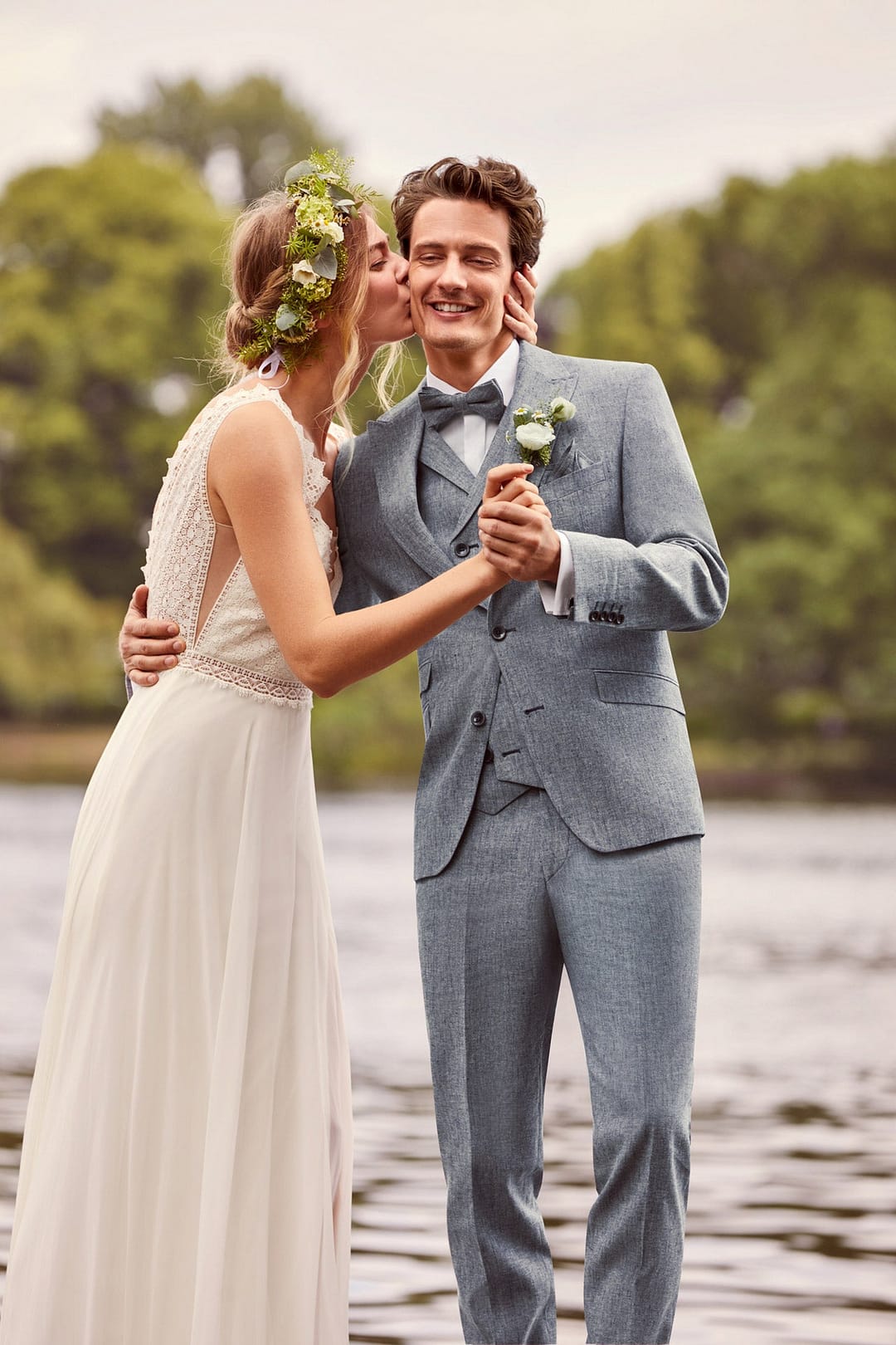 Eine Braut in weißem Kleid und Blumenkranz küsst ihren lächelnden Bräutigam, der die Atelier Torino Vintage Wedding Hose 811773/30 mit einem hellblauen Anzug trägt. Sie halten Händchen an einem See, umgeben von grünen Bäumen.