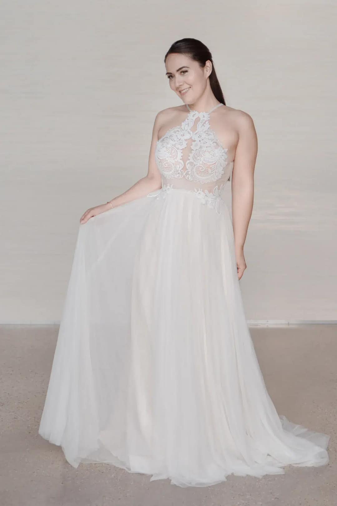 Eine Frau in dem ärmellosen, bodenlangen Bridonista C24-05 Hochzeitskleid lächelt, während sie einen Teil ihres Rocks hochhält. Ihr langes dunkles Haar ist zurückgesteckt, und sie steht vor einem schlichten, hellen Hintergrund.