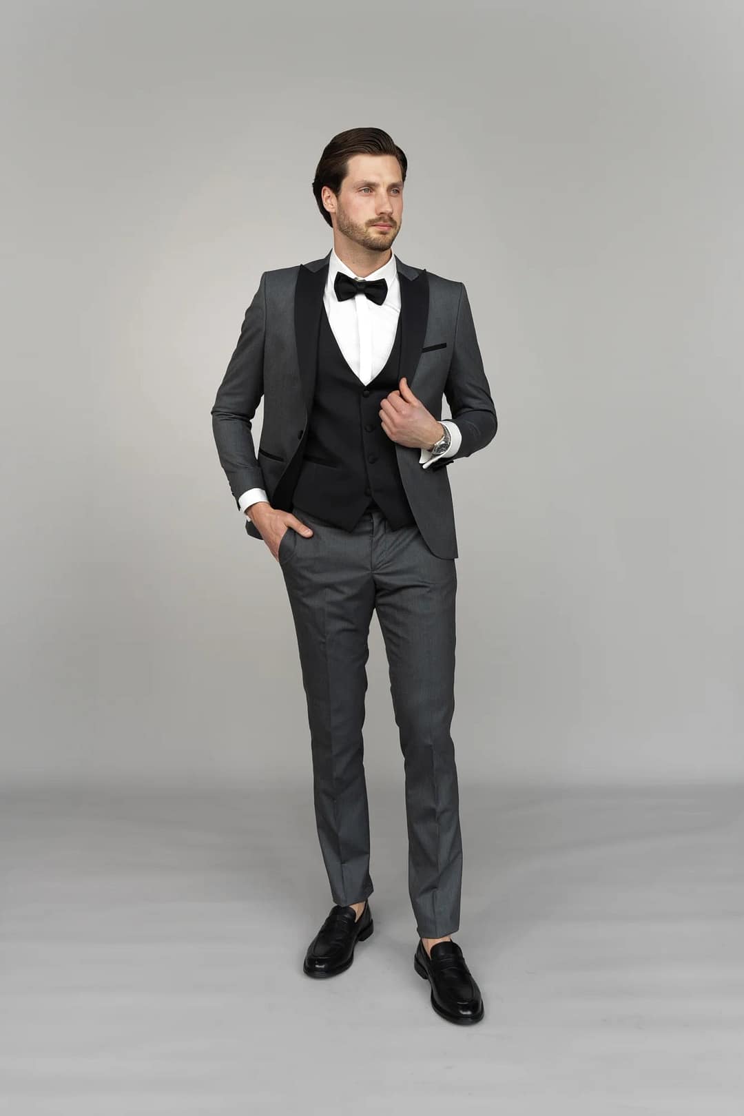Ein Mann steht vor einem schlichten grauen Hintergrund und trägt den Anzug Masculini 3397-08, eine stilvolle Wahl, die perfekt zu jedem Brautkleid oder Hochzeitskleid passt, mit einer Hand in der Tasche und einem Blick zur Seite.