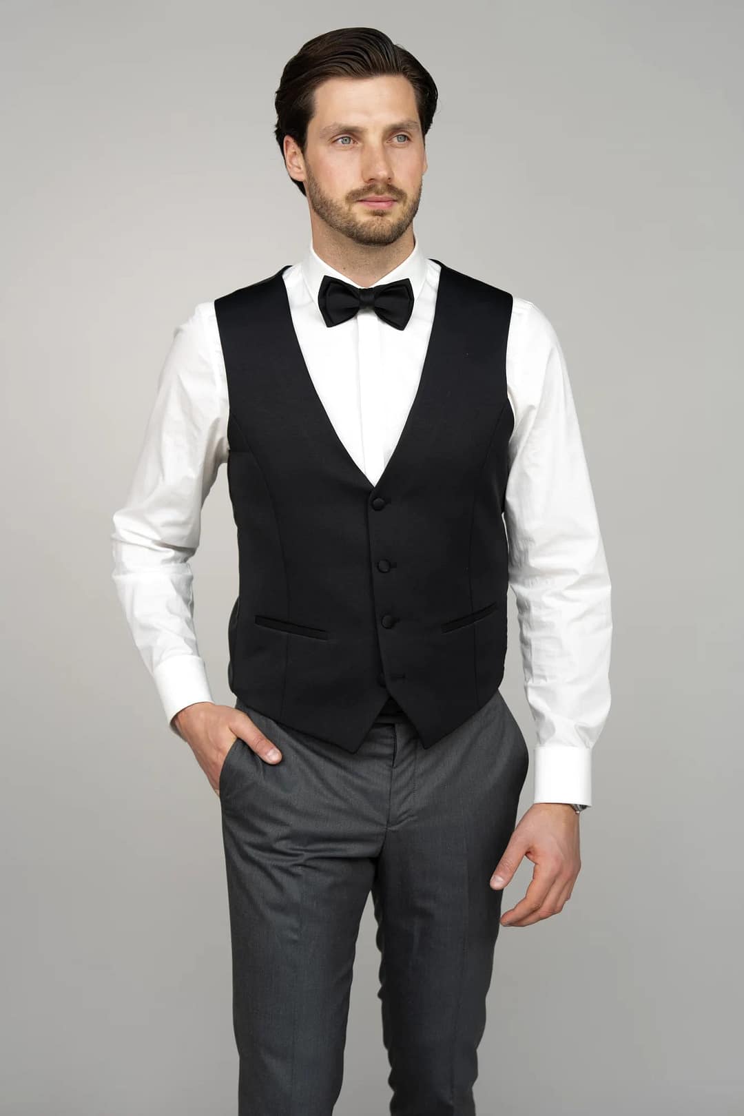 Ein Mann modelliert die Masculini 3397-08: schwarze Fliege, schwarze Weste, weißes Hemd und graue Hose - die perfekte formale Inspiration für Brautmoden oder Hochzeitskleider vor einem schlichten hellgrauen Hintergrund.