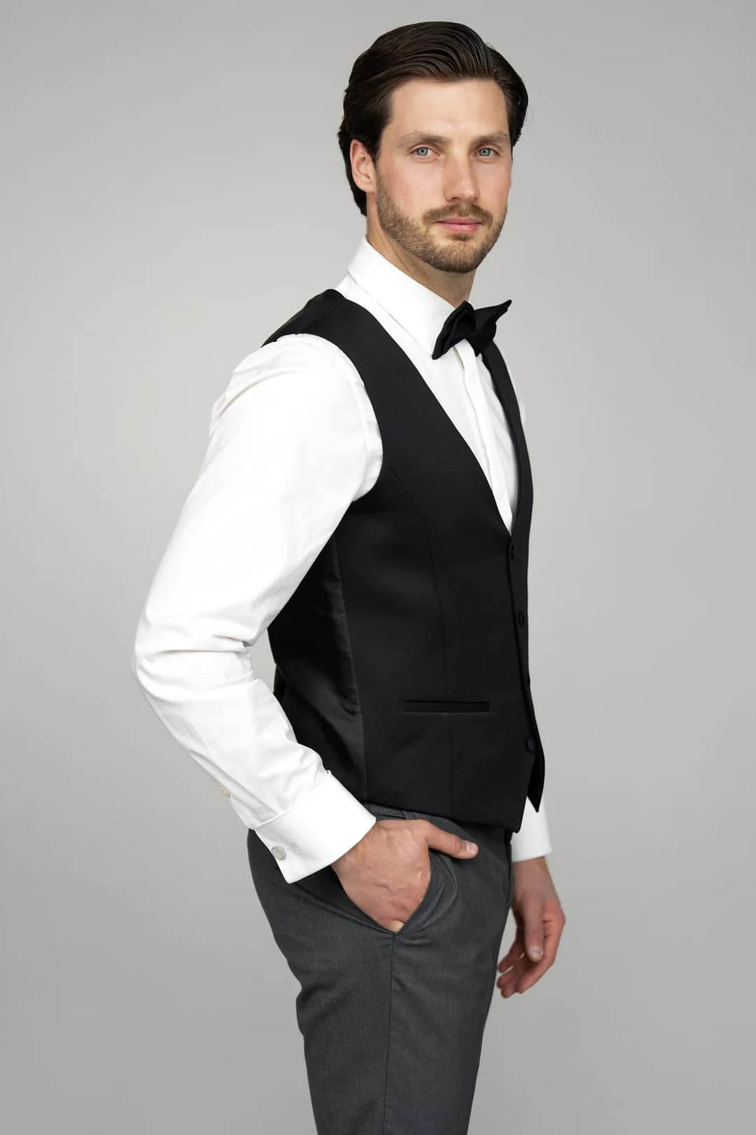 Ein Mann mit kurzen dunklen Haaren und Bart trägt das Modell Masculini 3397-08: ein weißes Hemd, eine schwarze Fliege, eine schwarze Weste und eine graue Hose. Er posiert seitwärts mit einer Hand in der Tasche vor einem schlichten grauen Hintergrund.