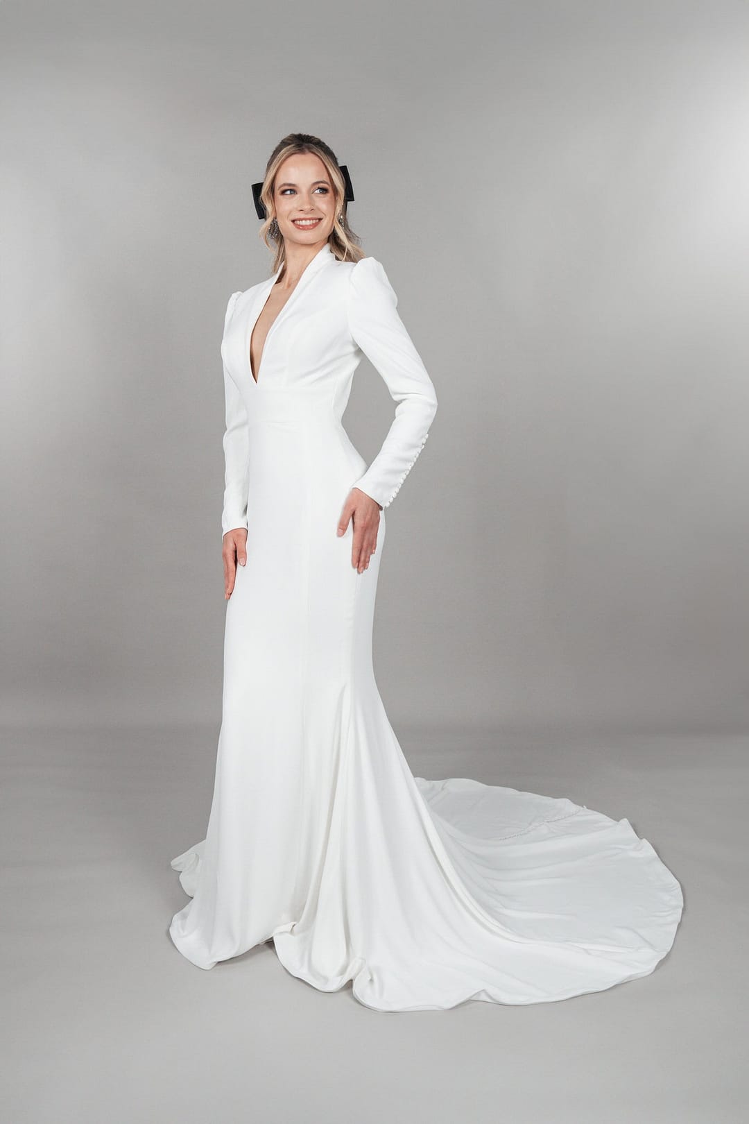 BC25-F09_HQ_6 Eine Frau steht lächelnd in dem Bridonista BC25-F09, einem langärmeligen, weißen, bodenlangen Brautkleid mit tailliertem Oberteil und leichter Schleppe, vor einem schlichten grauen Hintergrund.