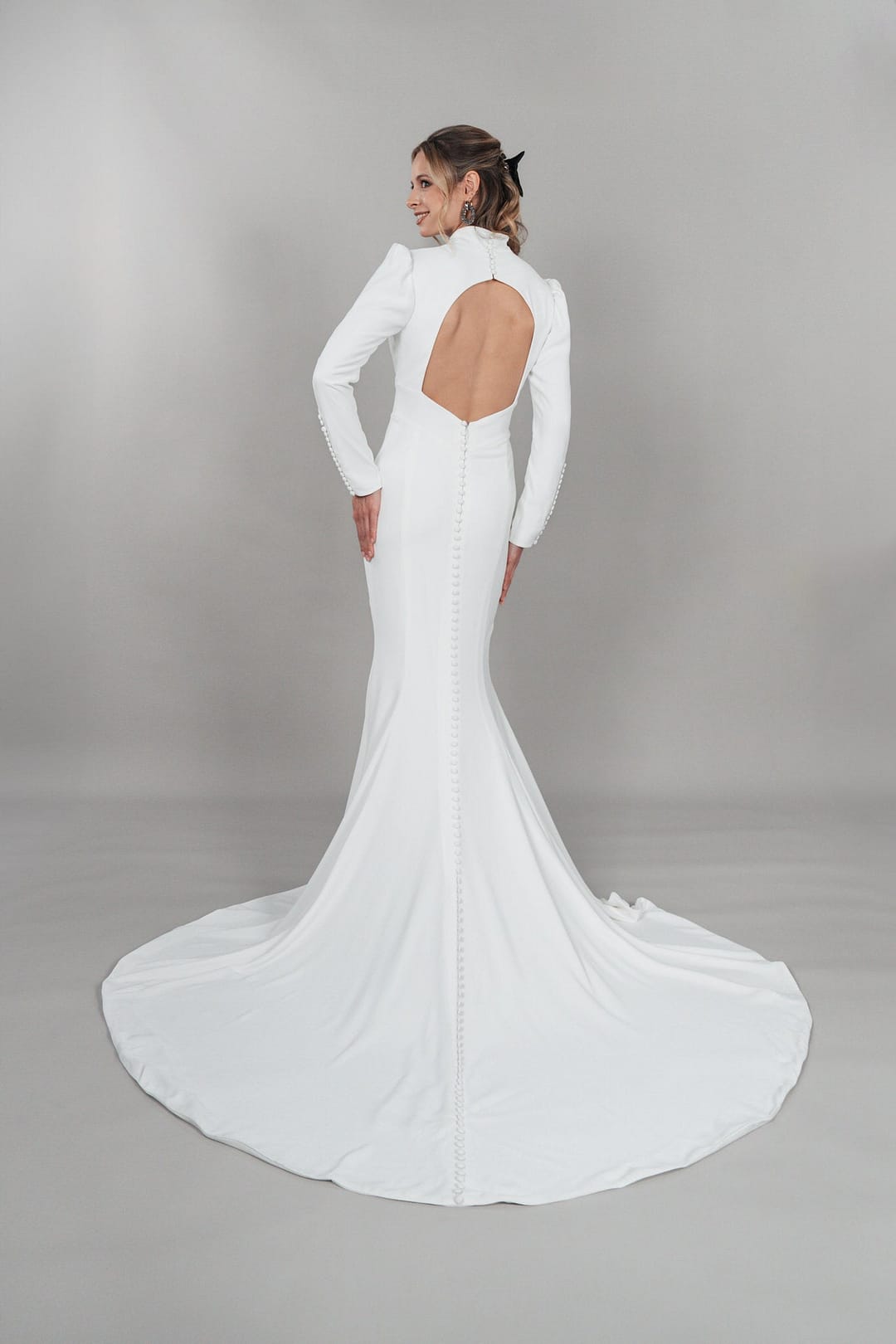 BC25-F09_HQ_8 Eine Frau steht vor einem grauen Hintergrund und trägt das Bridonista BC25-F09: ein langes, tailliertes weißes Brautkleid mit langen Ärmeln, einem offenen Rücken, einer Schleppe und einer Reihe von Knöpfen am Rücken.