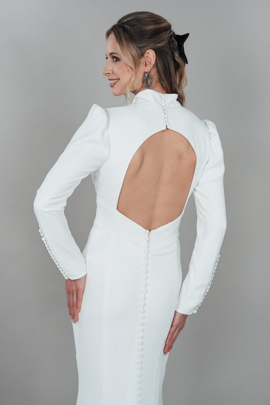 BC25-F09_HQ_9 Eine Frau trägt das Bridonista BC25-F09, ein langärmeliges weißes Hochzeitskleid mit offenem Rücken, Knopfleiste und hohem Kragen. Sie steht vor einem schlichten grauen Hintergrund und lächelt über ihre Schulter.