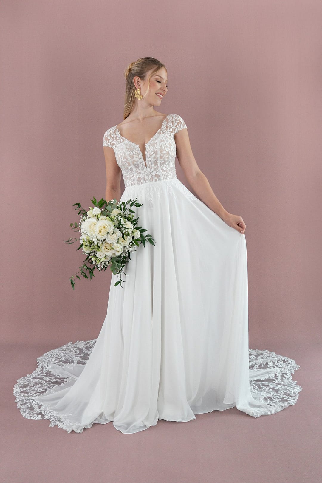 V26-A05_Web_1 Eine lächelnde Braut im Bridonista V26-A05 Brautkleid hält einen weißen Blumenstrauß und steht vor einem zartrosa Hintergrund. Die Schleppe ihres eleganten Hochzeitskleides breitet sich anmutig hinter ihr aus.