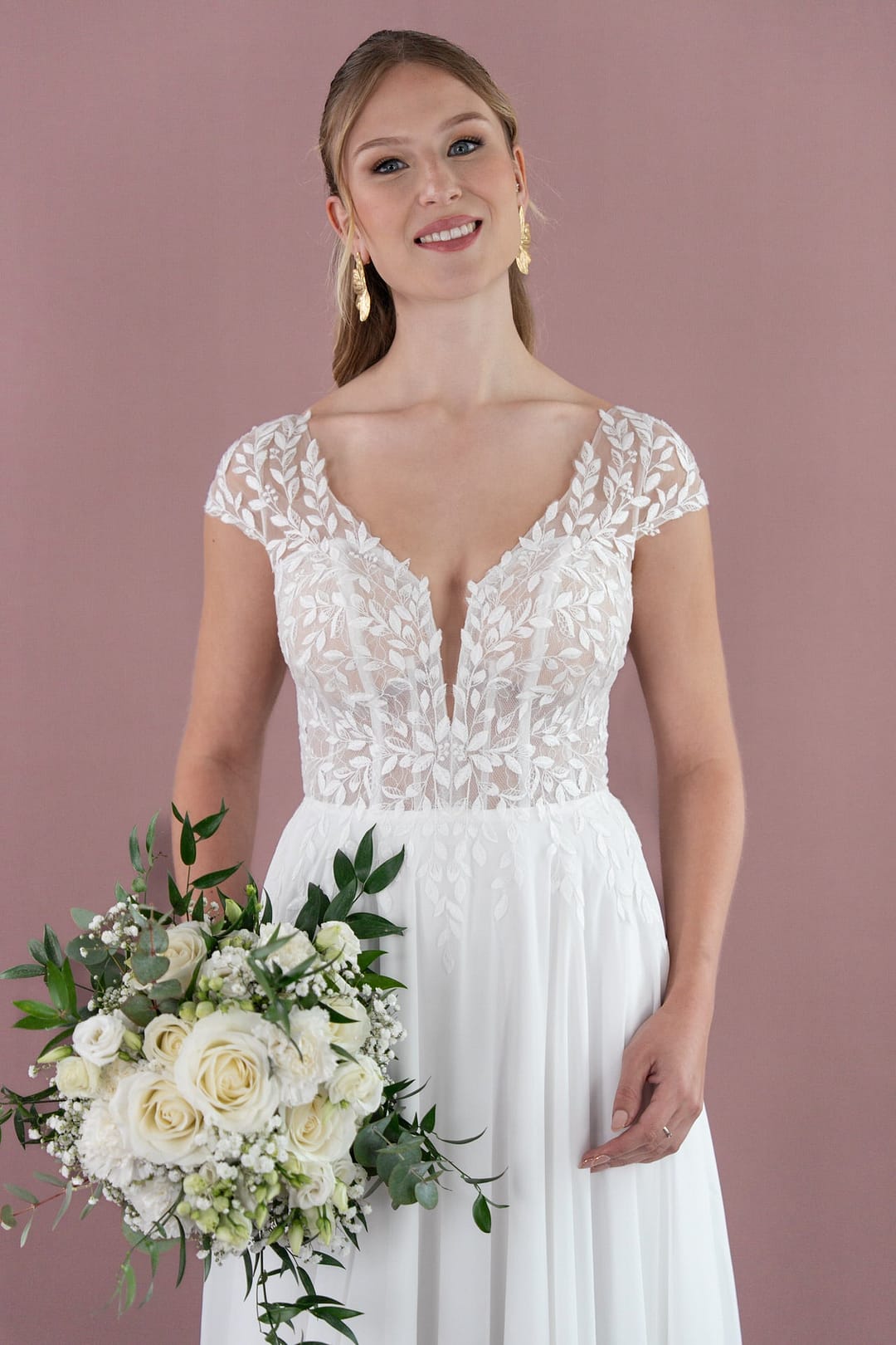 V26-A05_Web_2 Eine Frau, die die Bridonista V26-A05 trägt, lächelt sanft in die Kamera und hält einen Strauß weißer Rosen und Grünpflanzen vor einem schlichten lila Hintergrund.