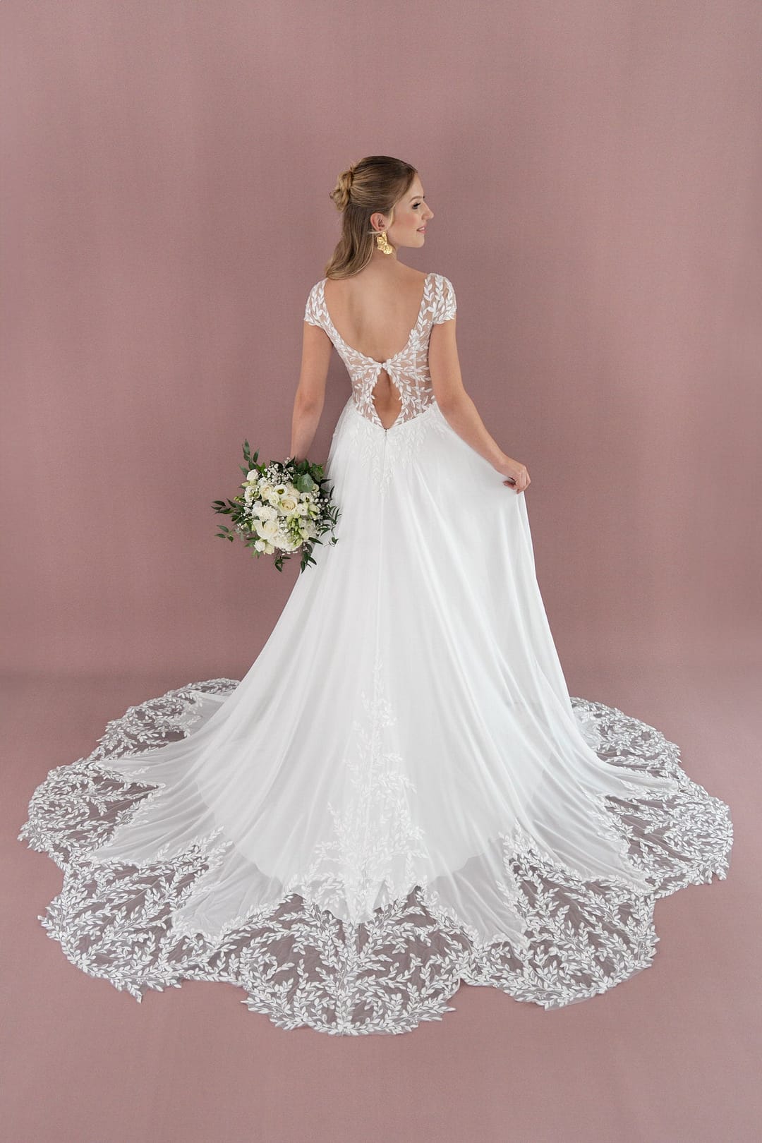 V26-A05_Web_3 Eine Braut in einem weißen Bridonista V26-A05 Brautkleid mit Spitzenverzierung und langer Schleppe steht vor einem rosafarbenen Hintergrund, hält einen Blumenstrauß in der Hand und wendet sich ab, um die offene Rückseite ihres Hochzeitskleides zu zeigen.
