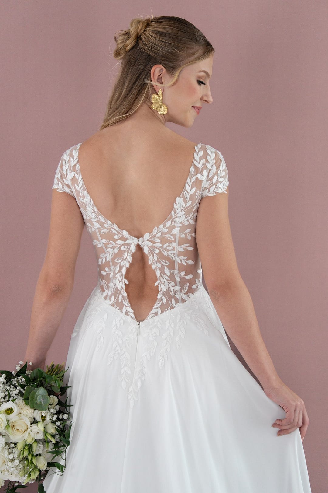 V26-A05_Web_5 Eine Frau im weißen Brautkleid Bridonista V26-A05 mit durchsichtigem, blattgemustertem Rücken und Kappenärmeln hält einen Strauß weißer Blumen. Sie trägt goldene Ohrringe und ihr Haar ist zu einer Hochsteckfrisur frisiert, die vor einem zartrosa Hintergrund steht.